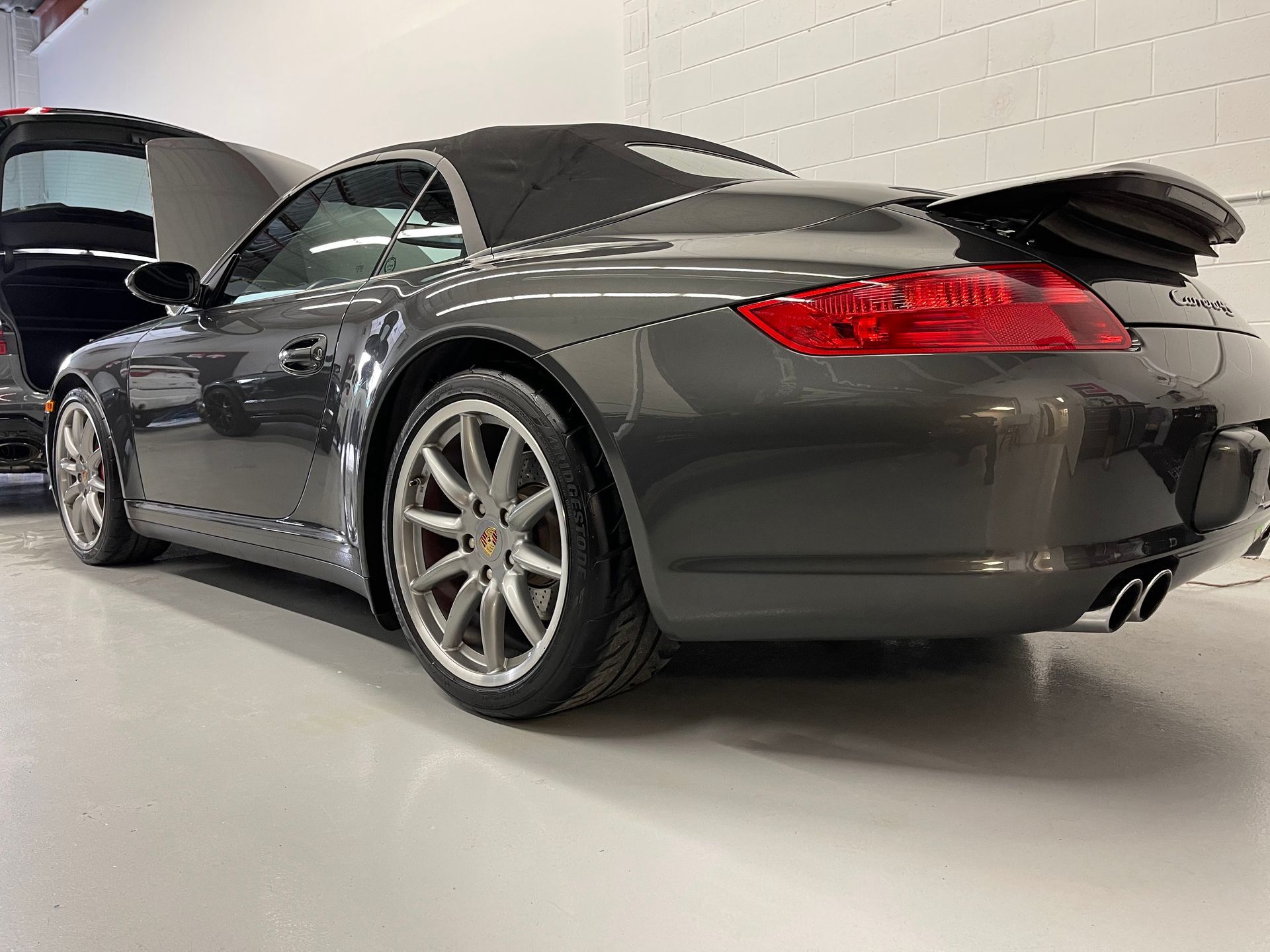 A dark gray Porsche convertible parked indoors on a glossy white floor, viewed from the rear three-quarter angle.