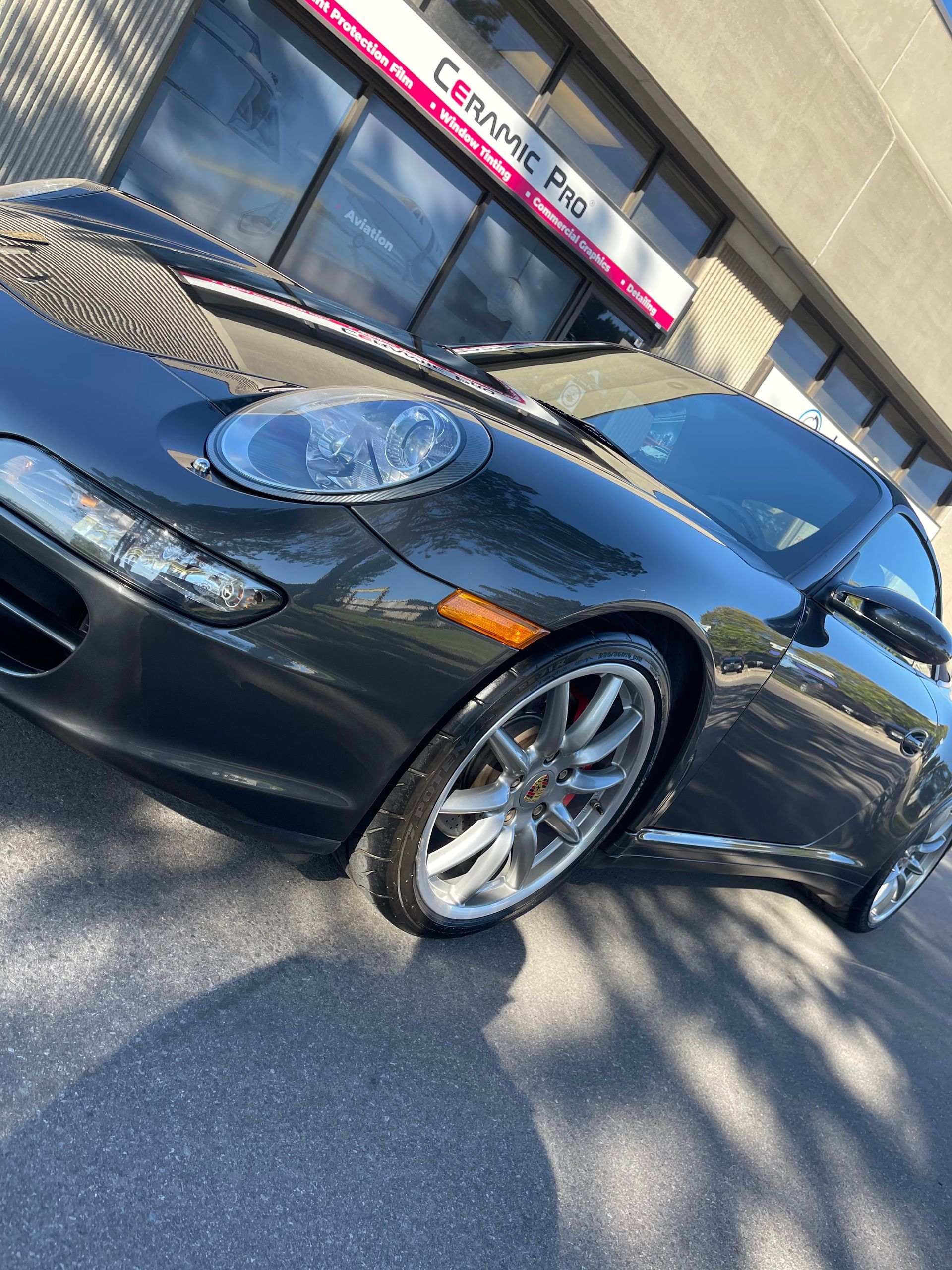 A dark grey Porsche convertible parked outside a Ceramic Pro shop on a sunny day.