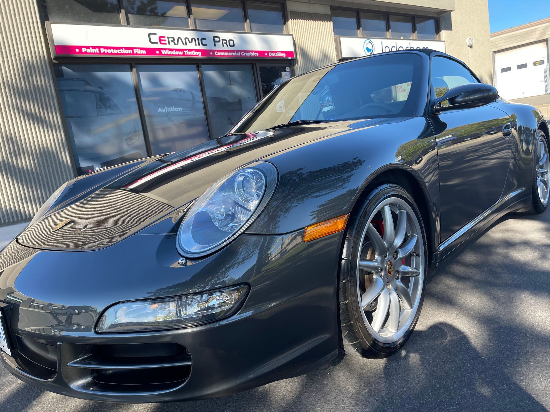 A dark gray Porsche 911 parked in front of a Ceramic Pro shop on a sunny day.