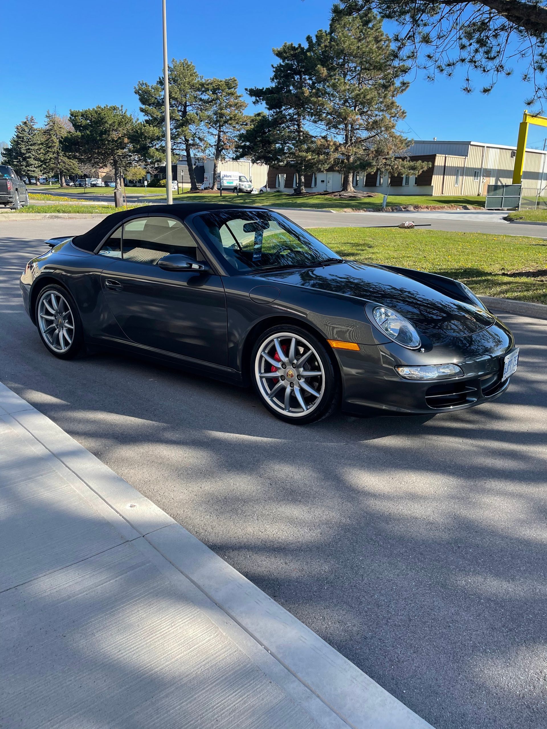 A dark gray Porsche convertible parked on an asphalt street next to a sidewalk with trees in the background.