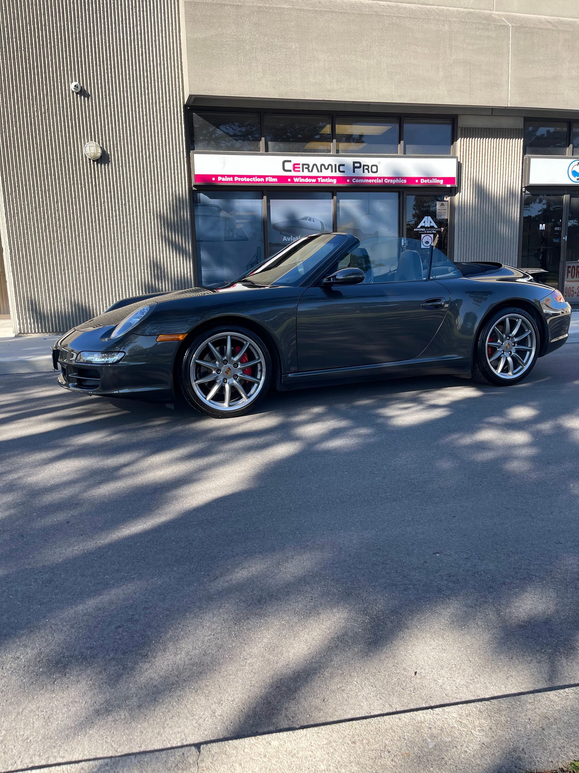 A dark grey Porsche 911 convertible parked in front of a commercial building on a sunny day.
