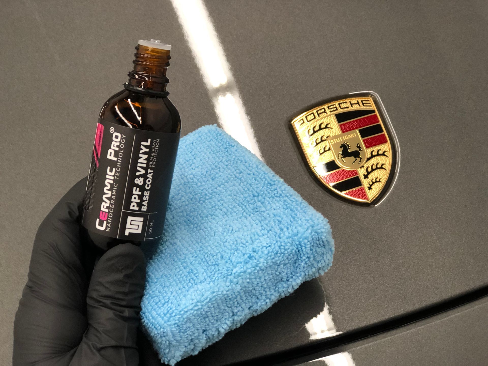 A gloved hand holds a bottle of Ceramic Pro PPF & Vinyl coating next to a blue applicator pad on a Porsche hood.