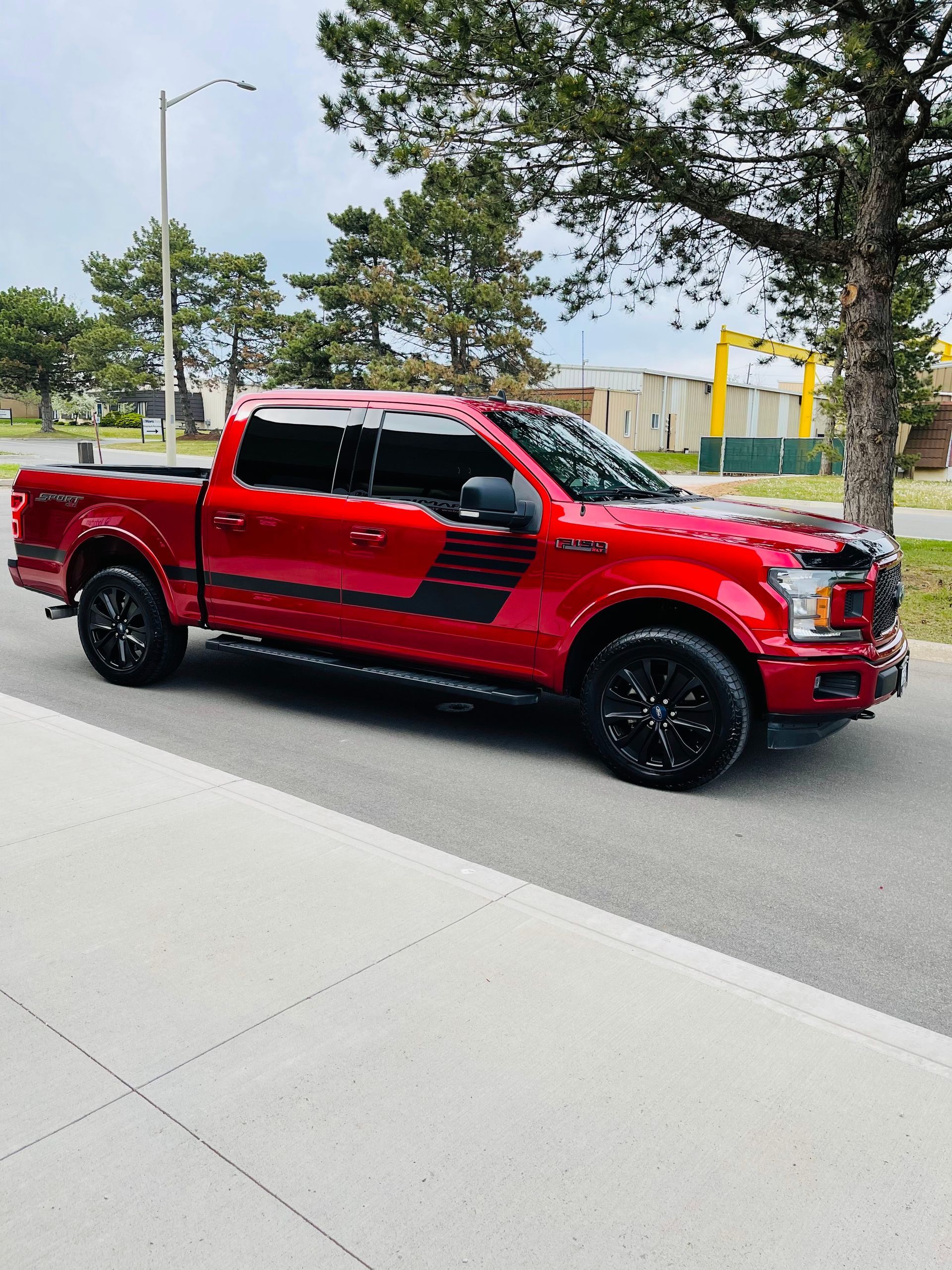 A red Ford F-150 pickup truck with black decals and wheels parked on an asphalt street near a tree.