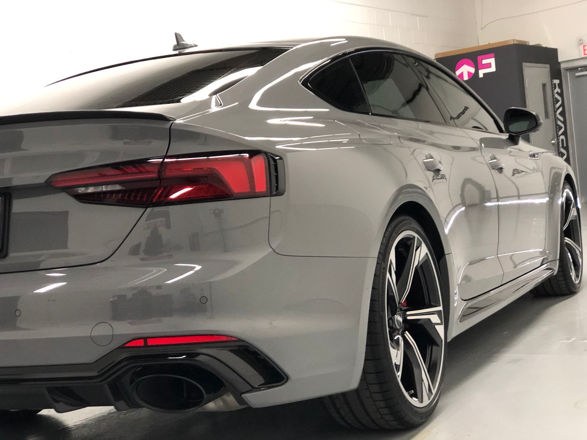 A sleek, grey Audi RS5 Sportback parked inside a brightly lit garage, showing its rear, taillights, and alloy wheels.