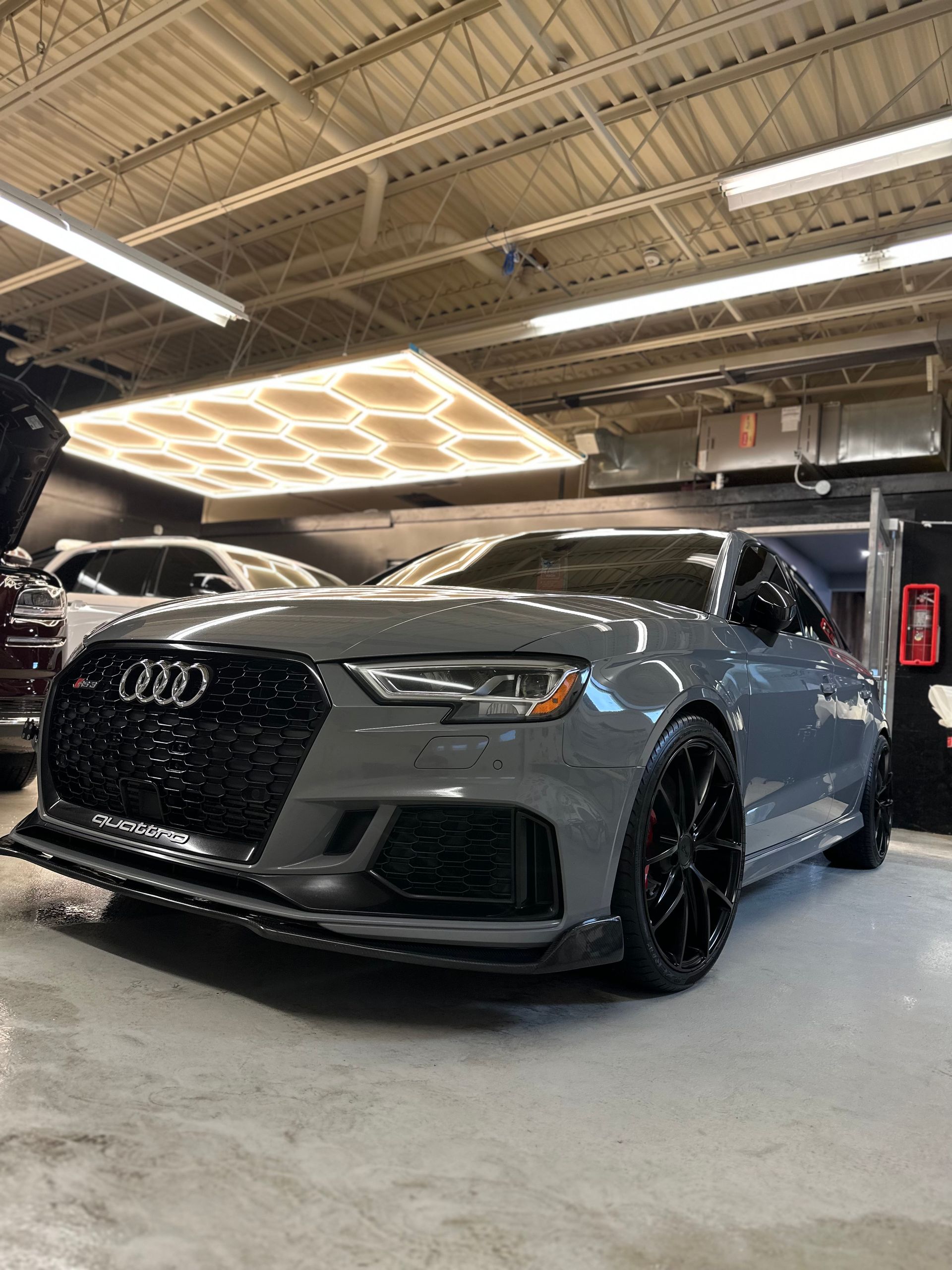 A Nardo gray Audi RS 3 with black wheels and a front splitter, parked inside a brightly lit garage.