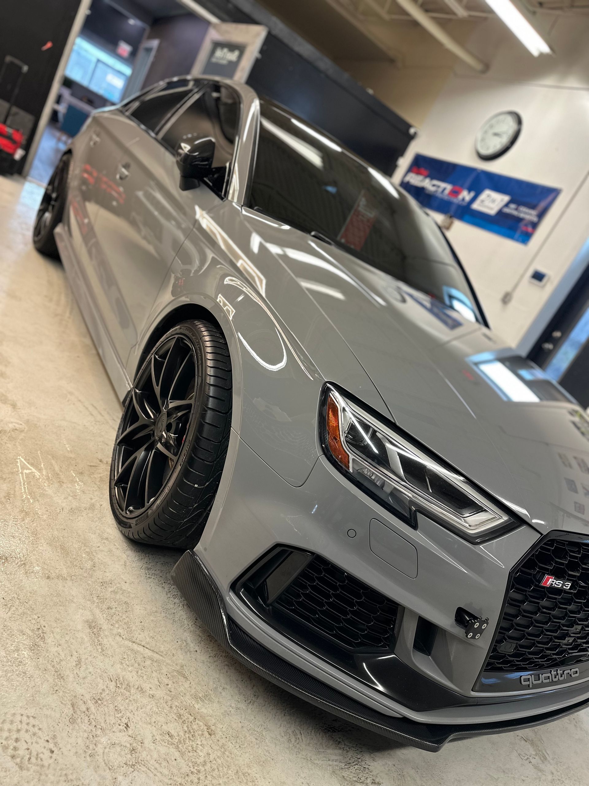 A nardo grey Audi RS3 sedan with black wheels and carbon fiber trim parked inside a garage.