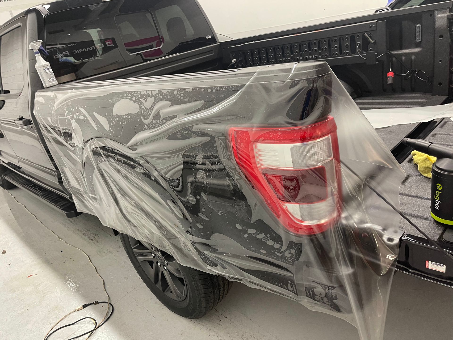 A grey truck bed panel covered in a translucent plastic protective film during an automotive detailing process.