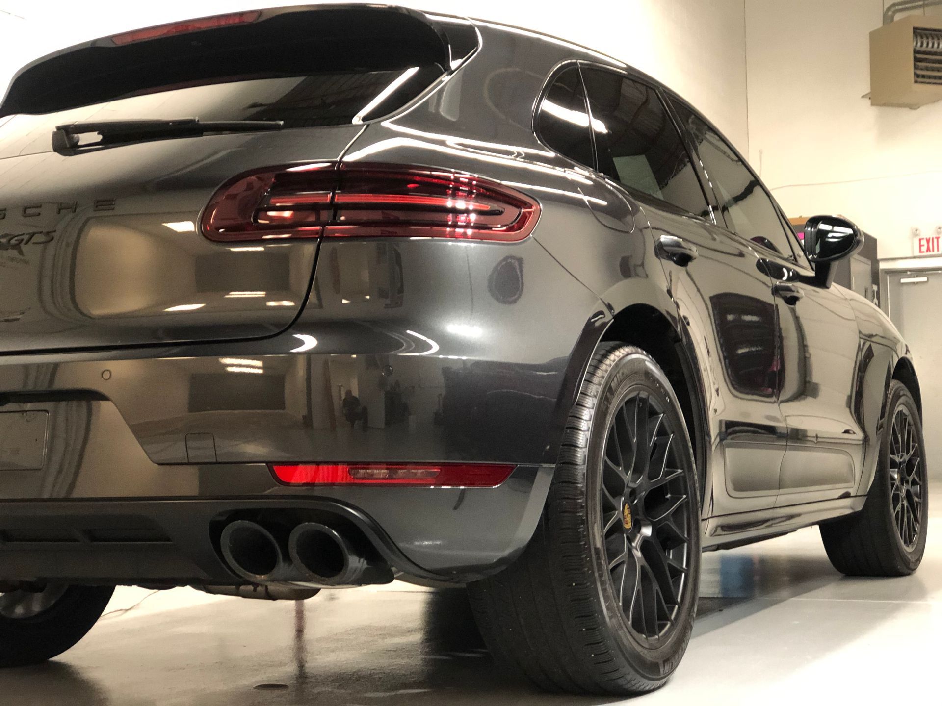 A grey Porsche Macan SUV shown from a low angle, parked indoors with black wheels and dual exhaust pipes.