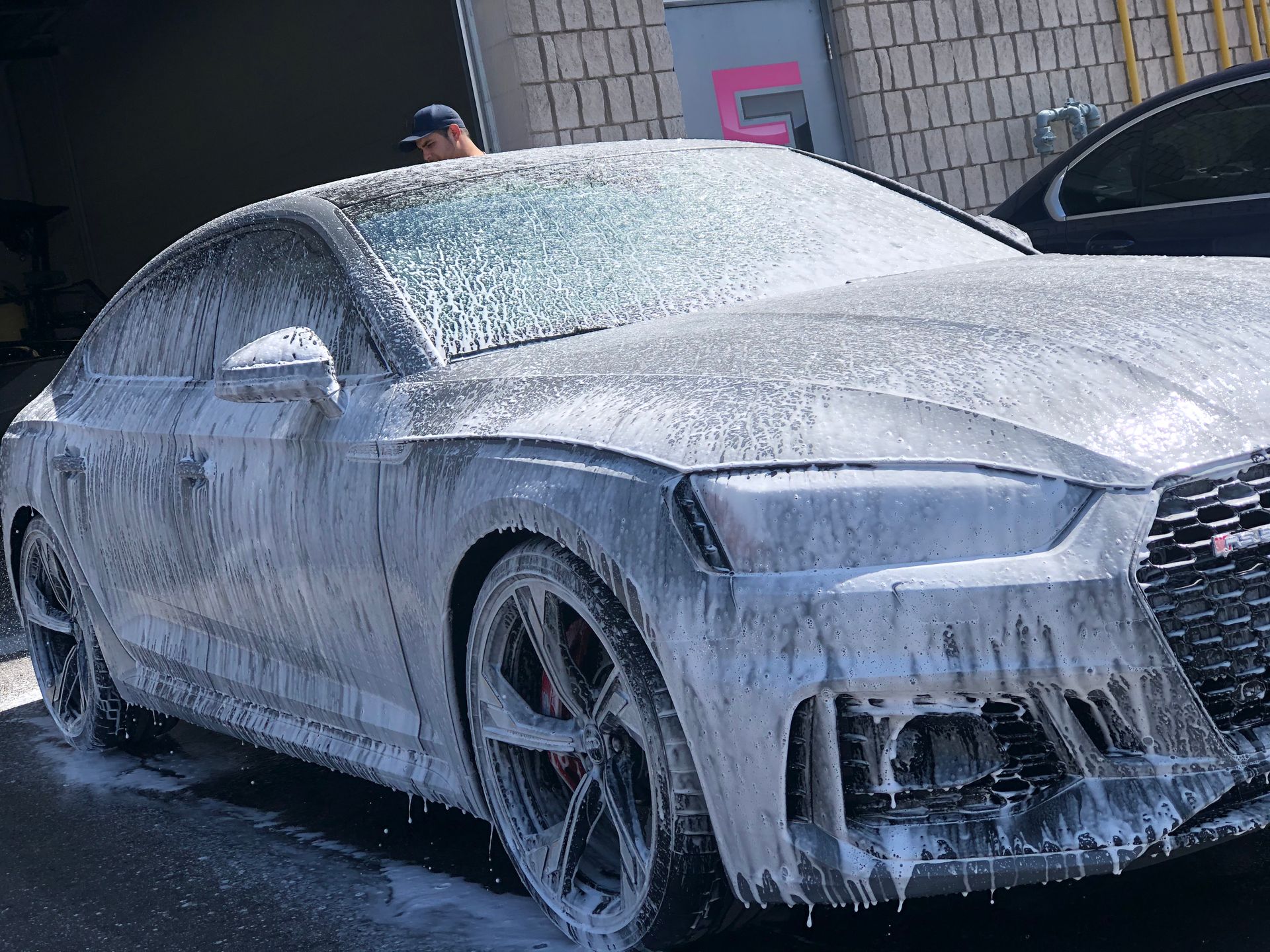 A luxury sedan covered in white sudsy cleaning foam during a car wash at a service bay.