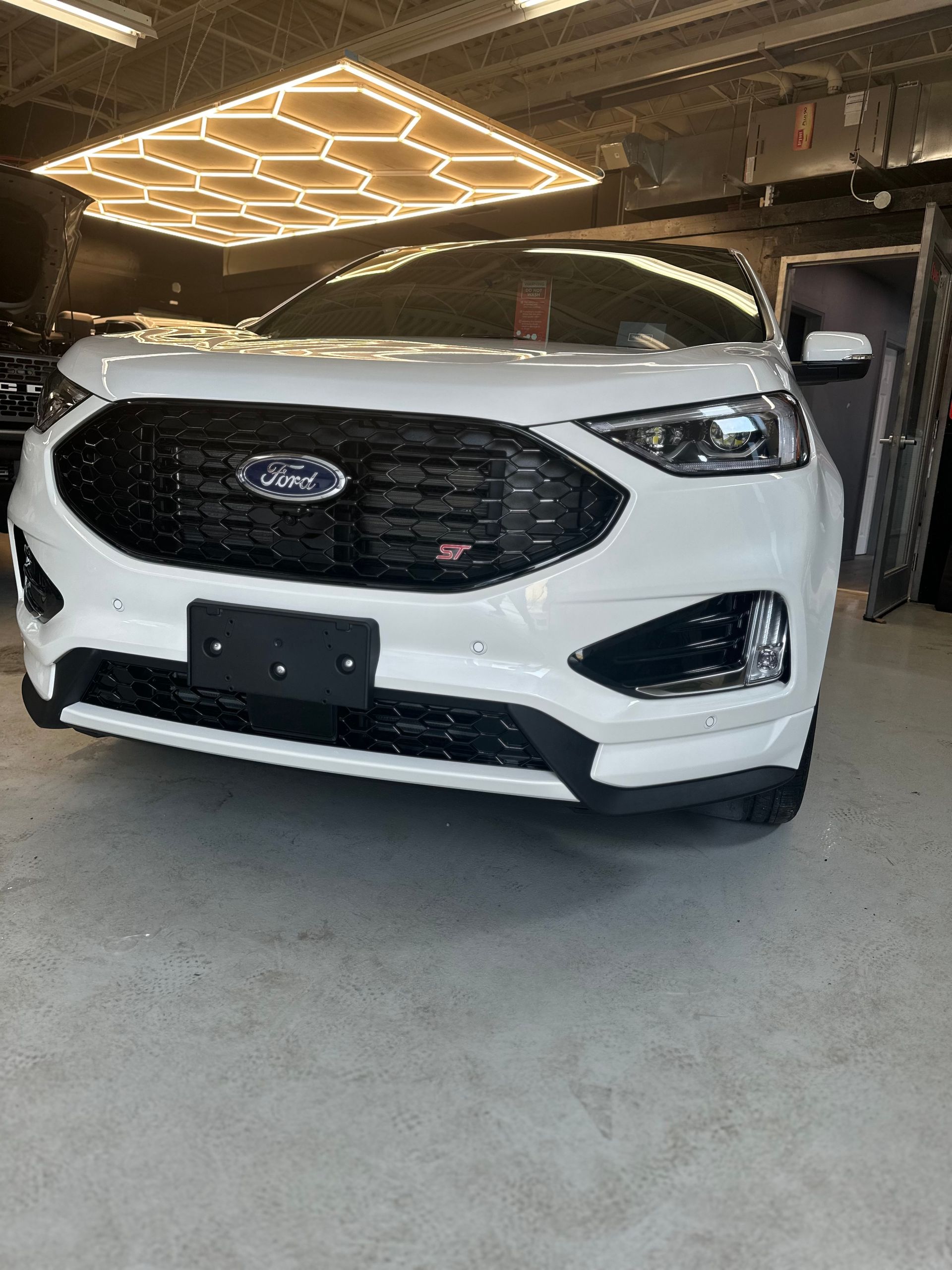 A white Ford Edge ST SUV parked in a garage with a hexagonal LED light fixture above.