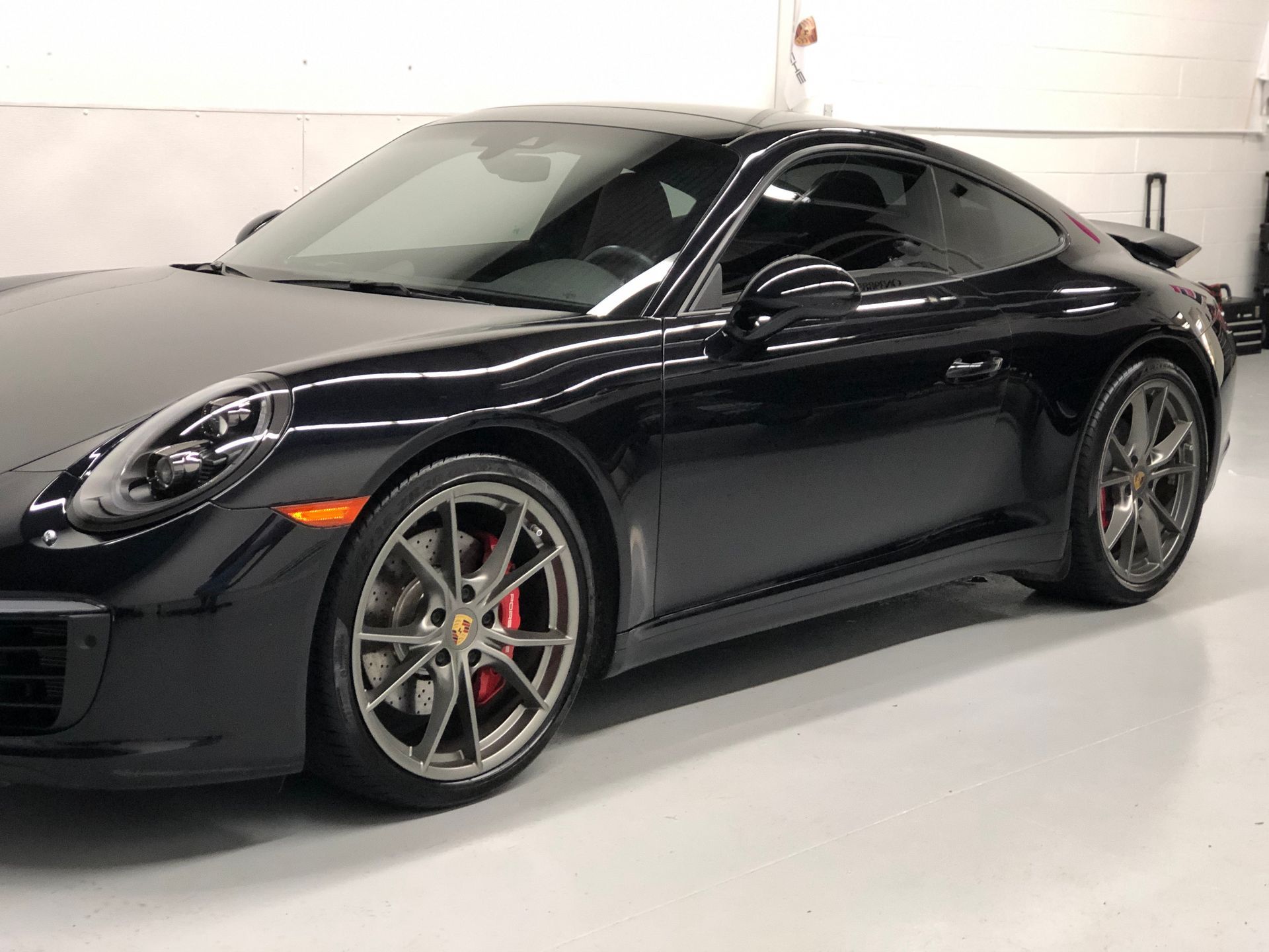 A black Porsche 911 coupe parked in a bright, clean garage with visible red brake calipers and grey alloy wheels.