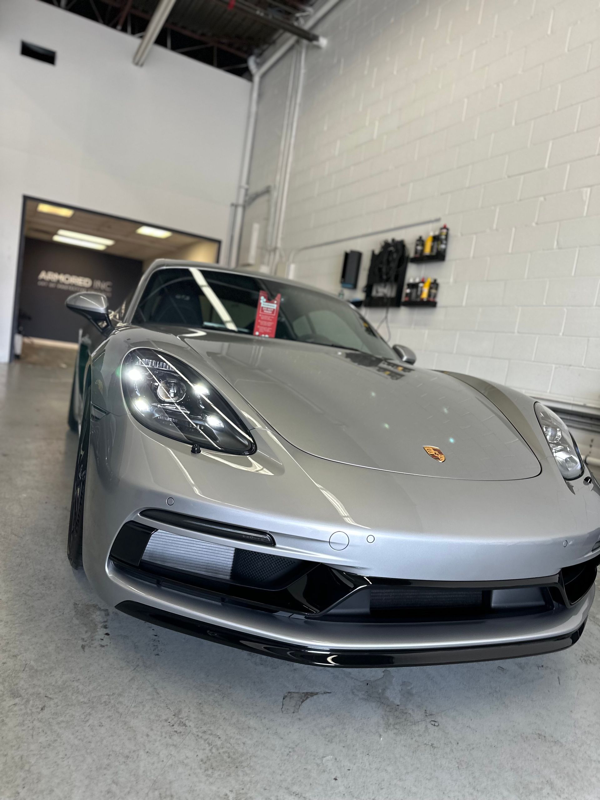 A silver Porsche parked inside an automotive detailing shop with bright lighting and white walls.