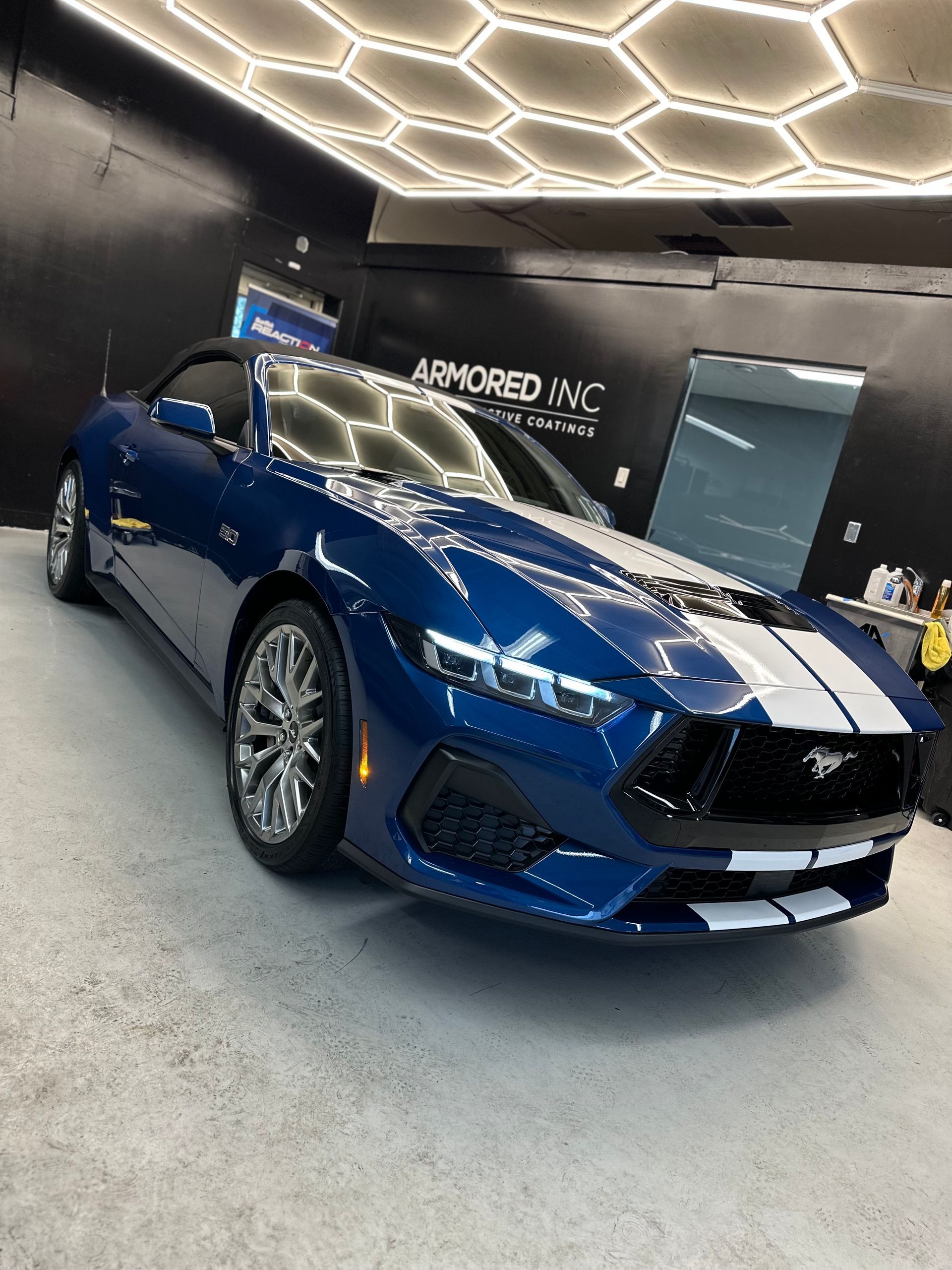 A blue Ford Mustang convertible with white racing stripes, parked in an indoor garage under hexagonal ceiling lights.