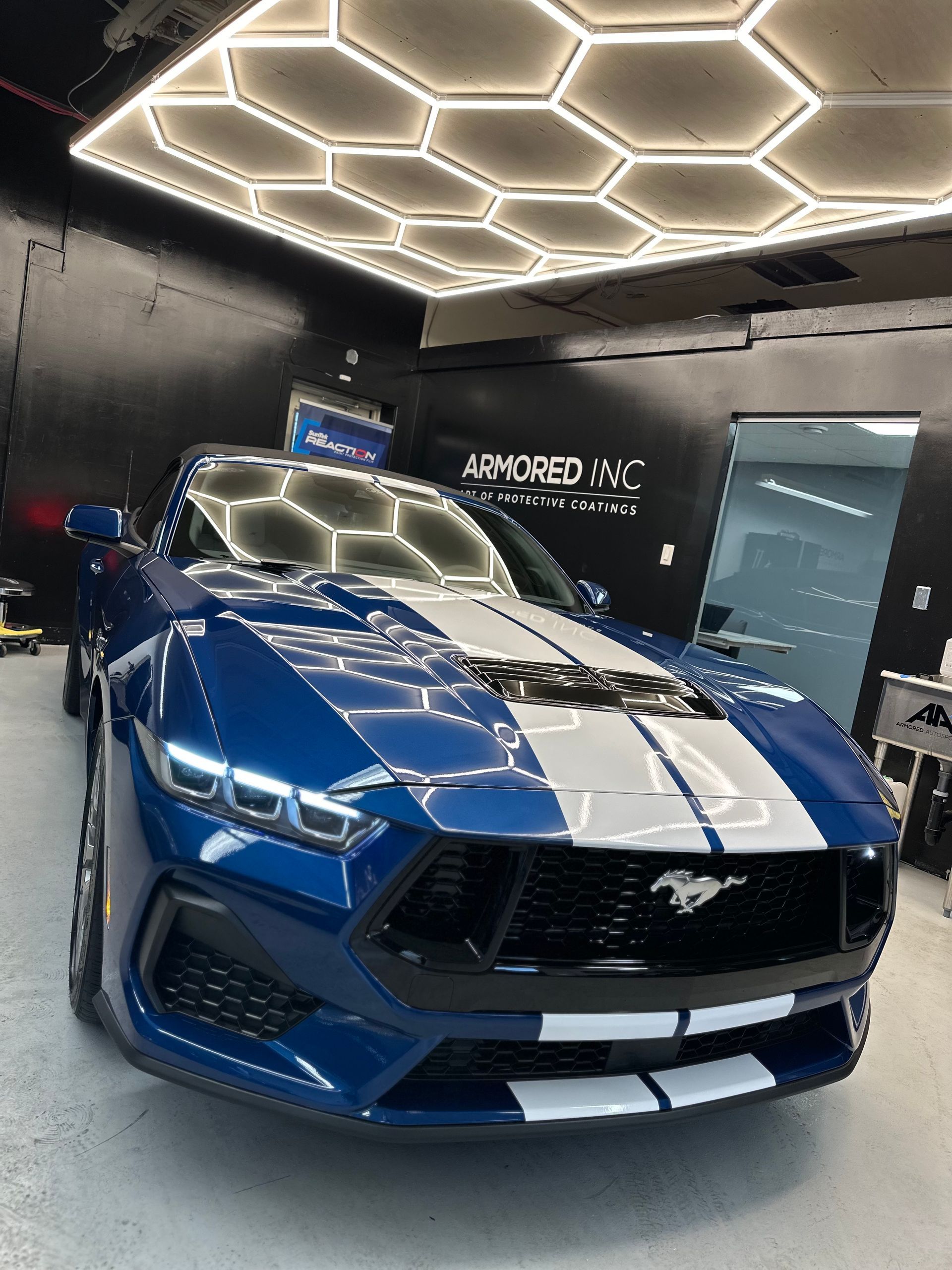 A blue Ford Mustang with white racing stripes parked inside a garage with hexagon-patterned overhead lighting.