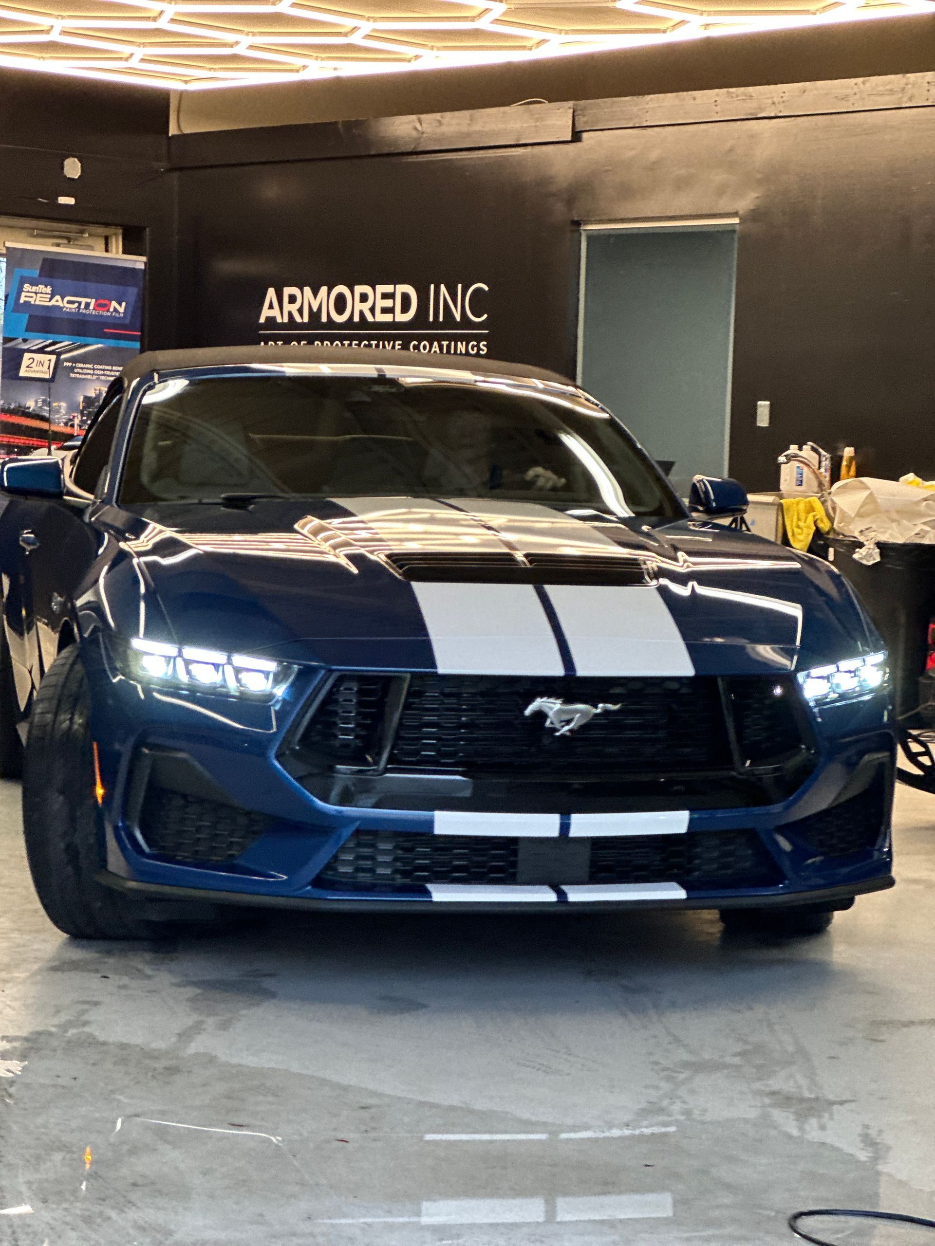 A dark blue Ford Mustang with white racing stripes parked inside an automotive shop called Armored Inc.