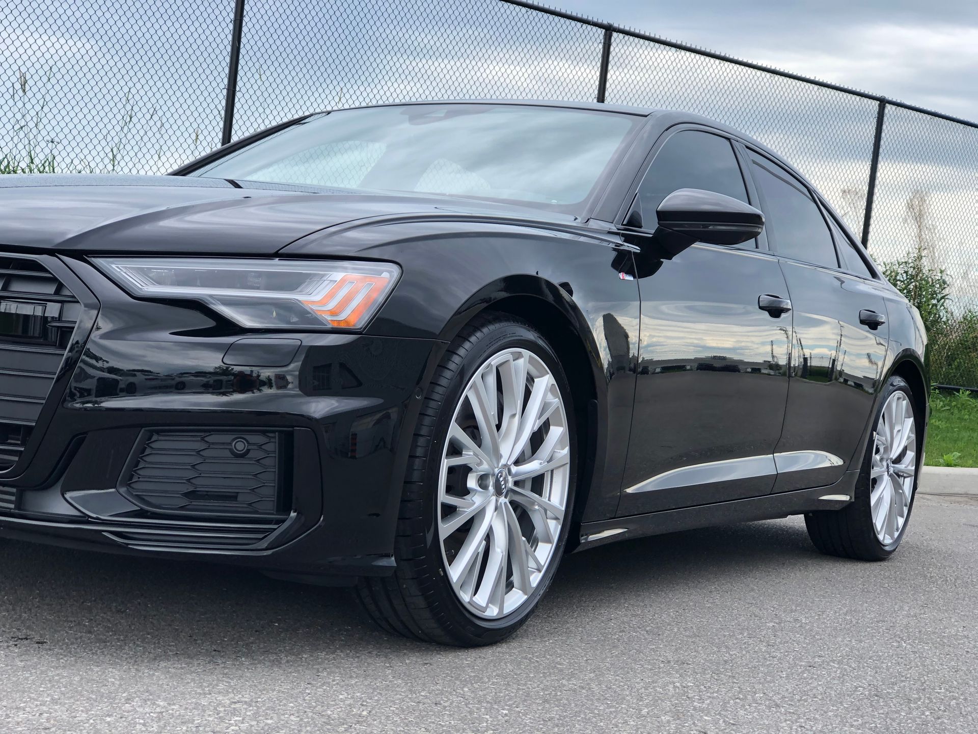A black Audi A6 sedan parked on asphalt in front of a chain-link fence.