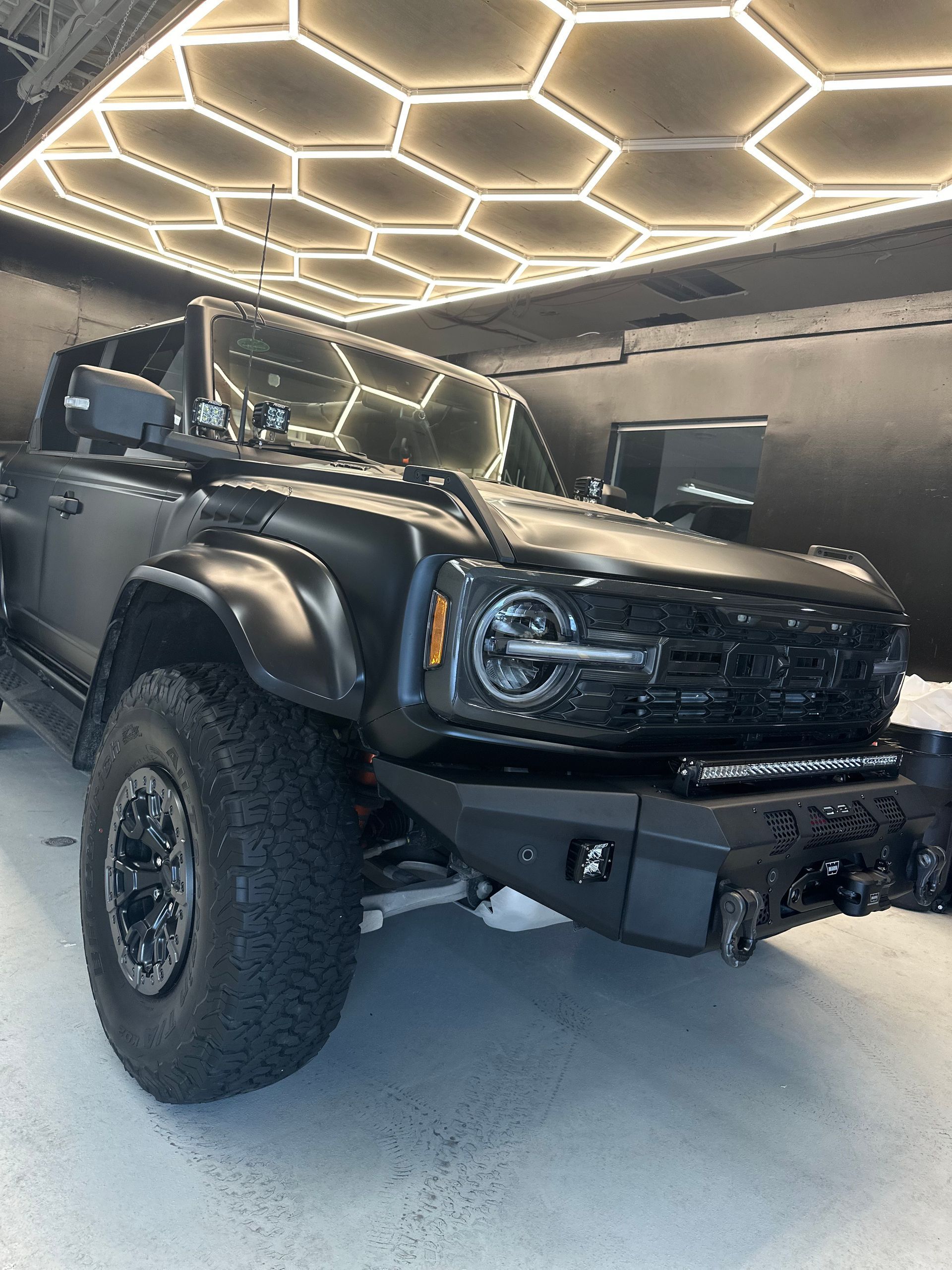 A black, modified Ford Bronco parked in a garage with distinctive hexagon-patterned ceiling lights.
