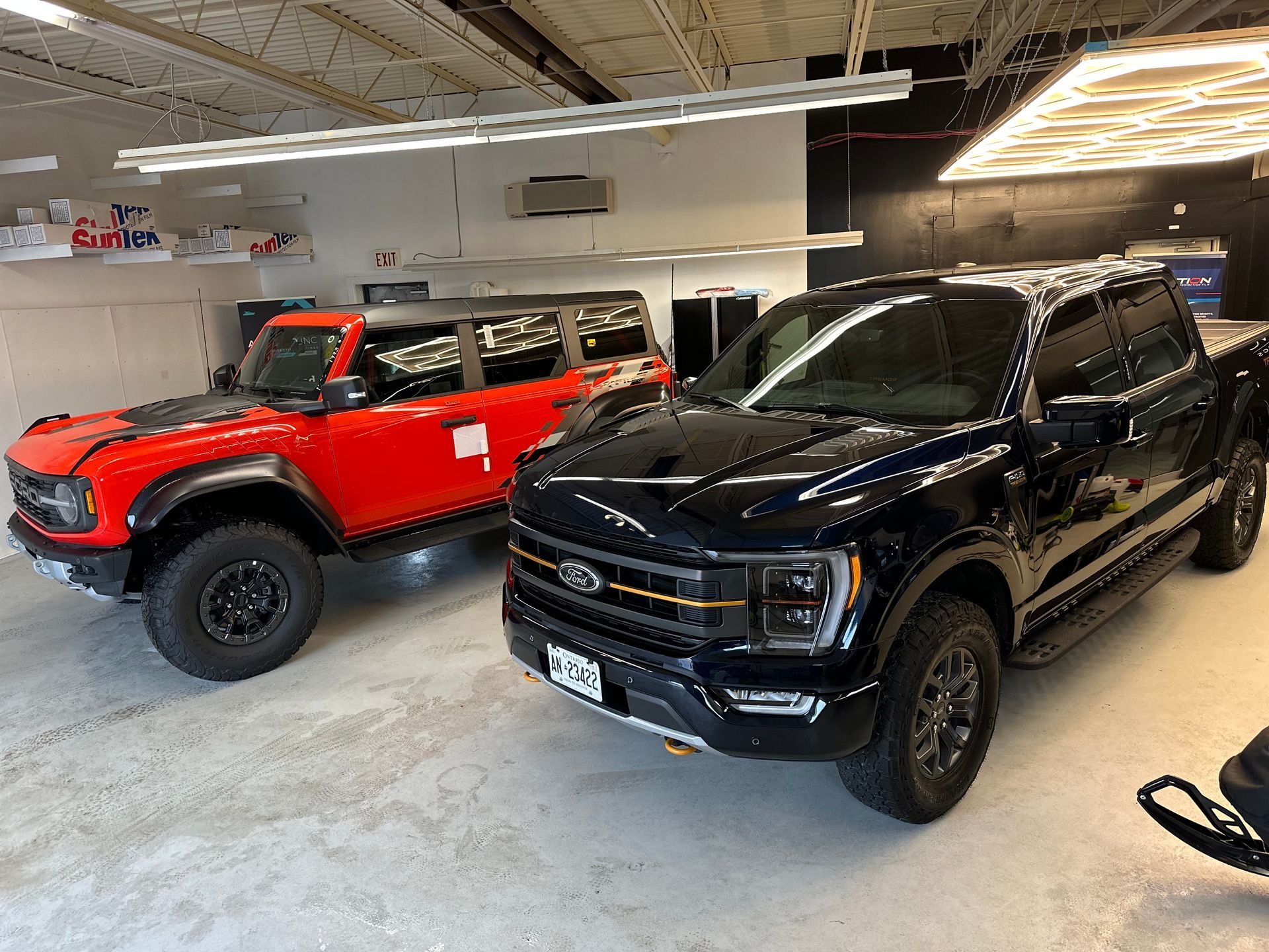 An orange Ford Bronco and a black Ford F-150 parked side-by-side in a well-lit indoor garage.