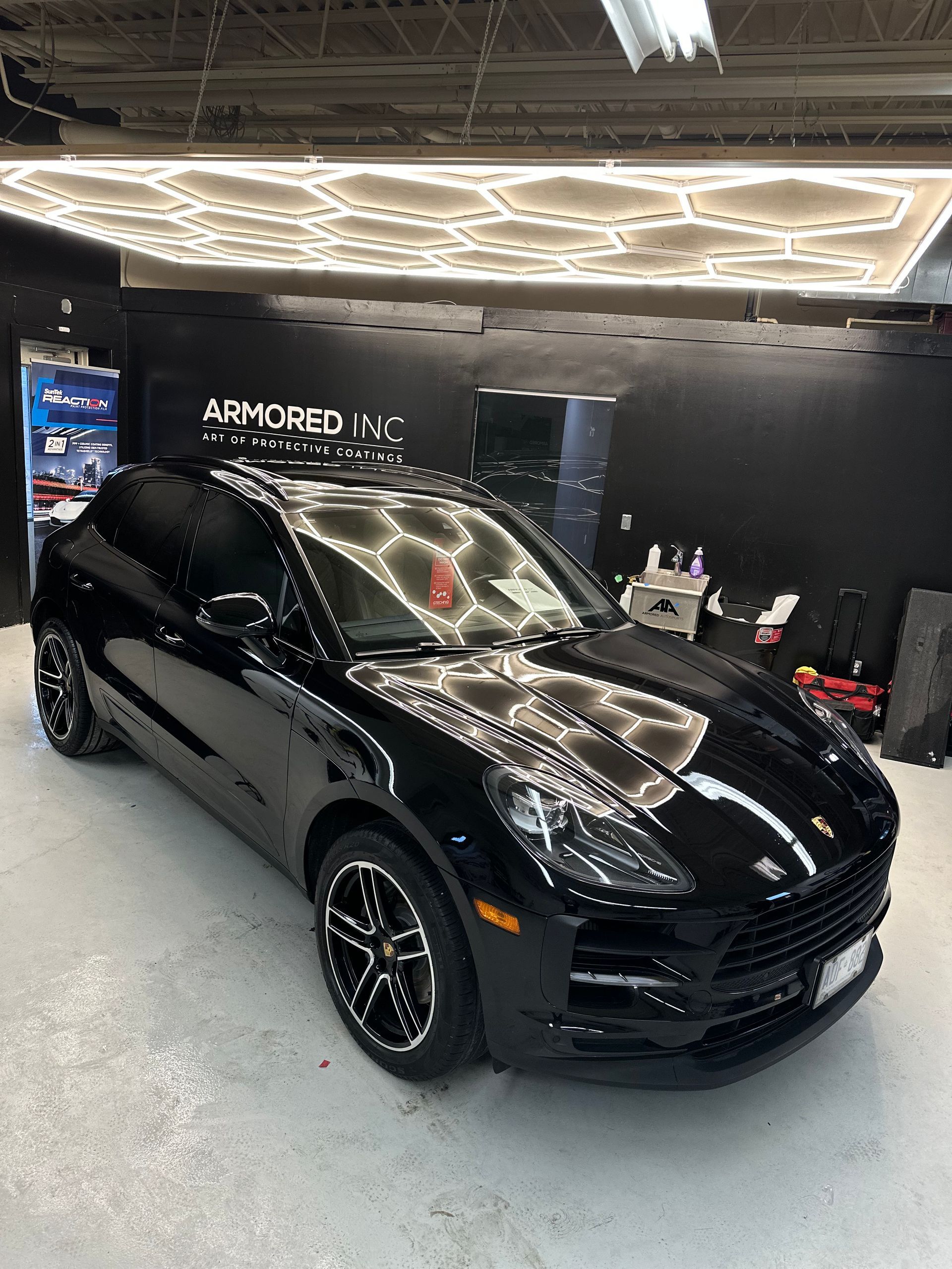 A shiny black Porsche SUV parked inside a garage with geometric ceiling lights and a black wall sign reading Armored Inc.