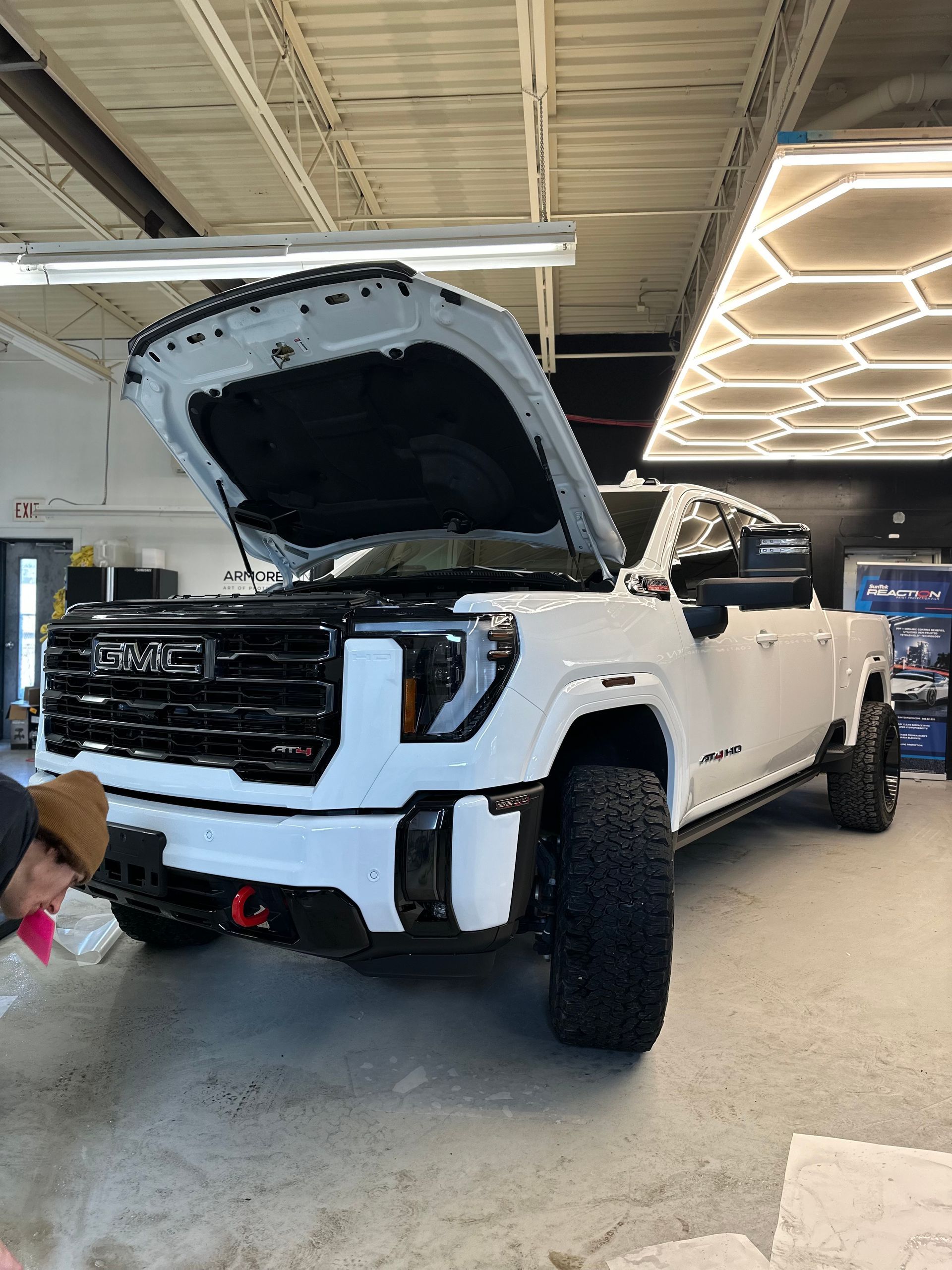 A white GMC pickup truck with its hood open, parked in an indoor garage with overhead hexagonal lighting.
