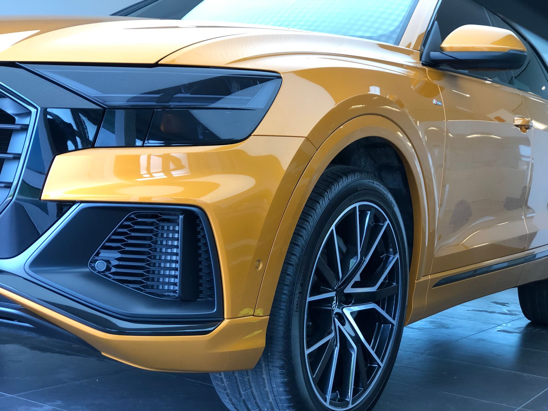 Close-up of the front corner of a gold Audi SUV, highlighting the headlight, black grille, and sporty alloy wheel.