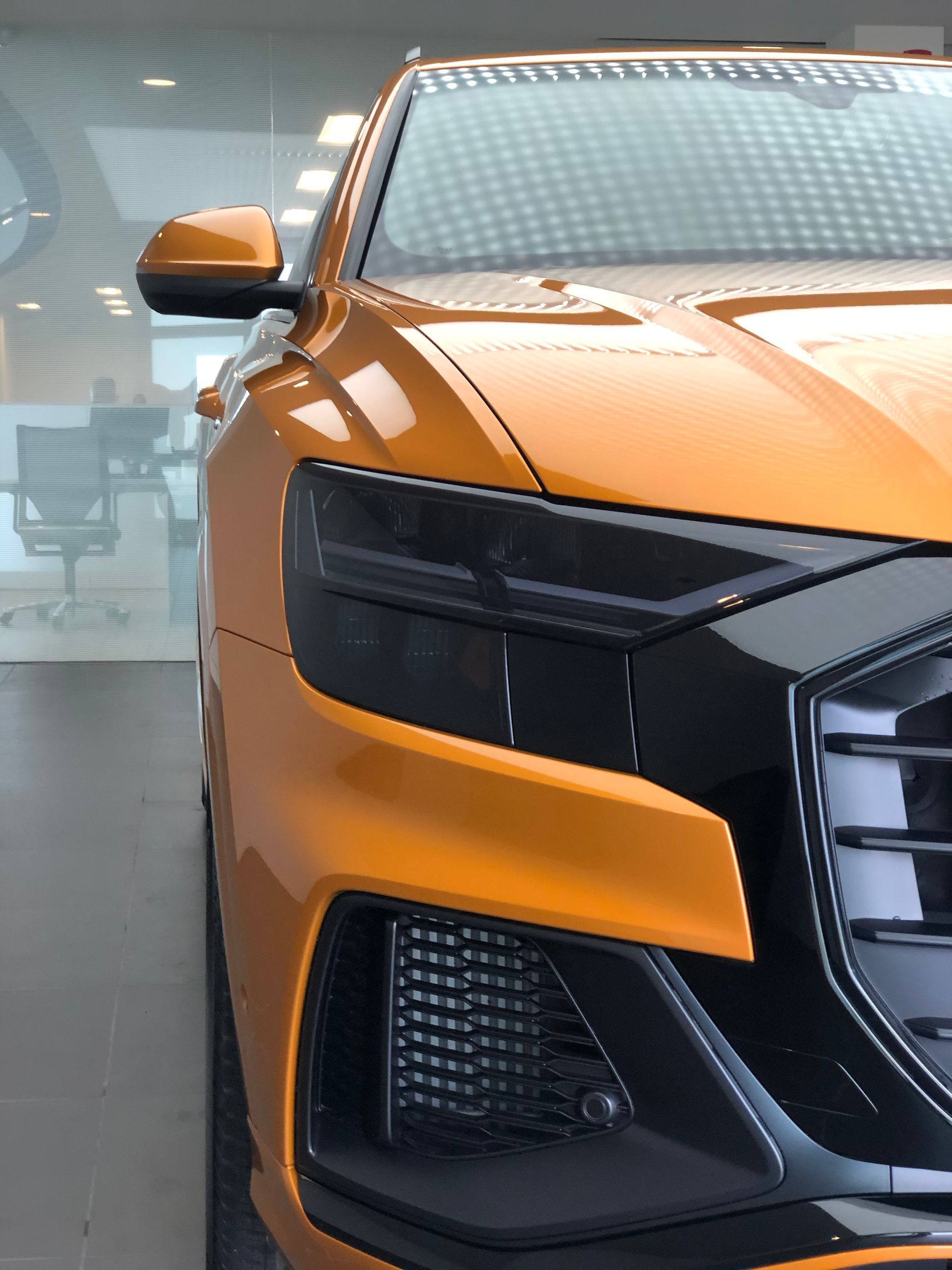 A close-up, angled view of the front corner of a bright orange luxury SUV, highlighting its sleek headlight and grille.