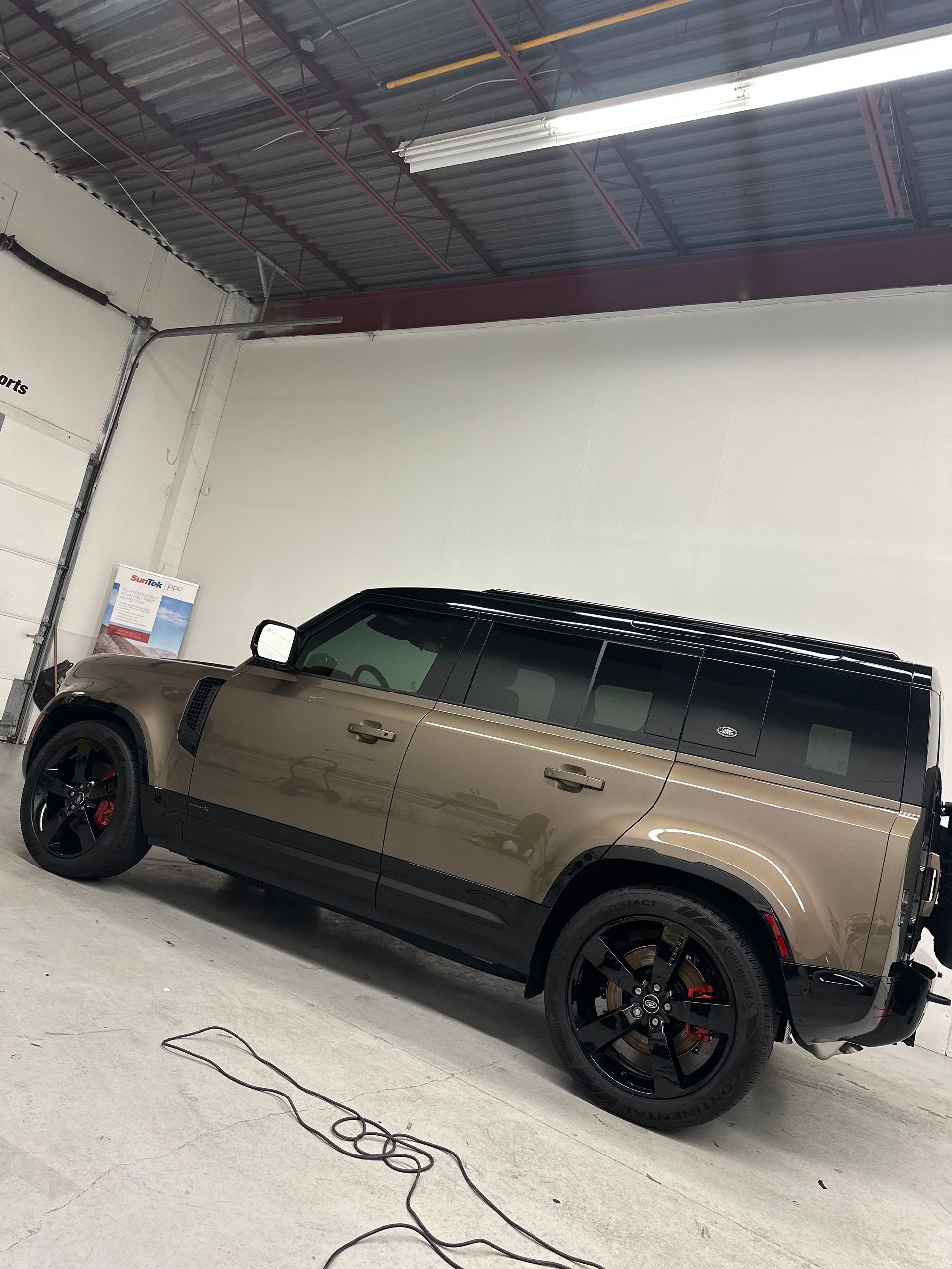 A side view of a metallic bronze Land Rover Defender with black rims parked in a bright, industrial warehouse space.