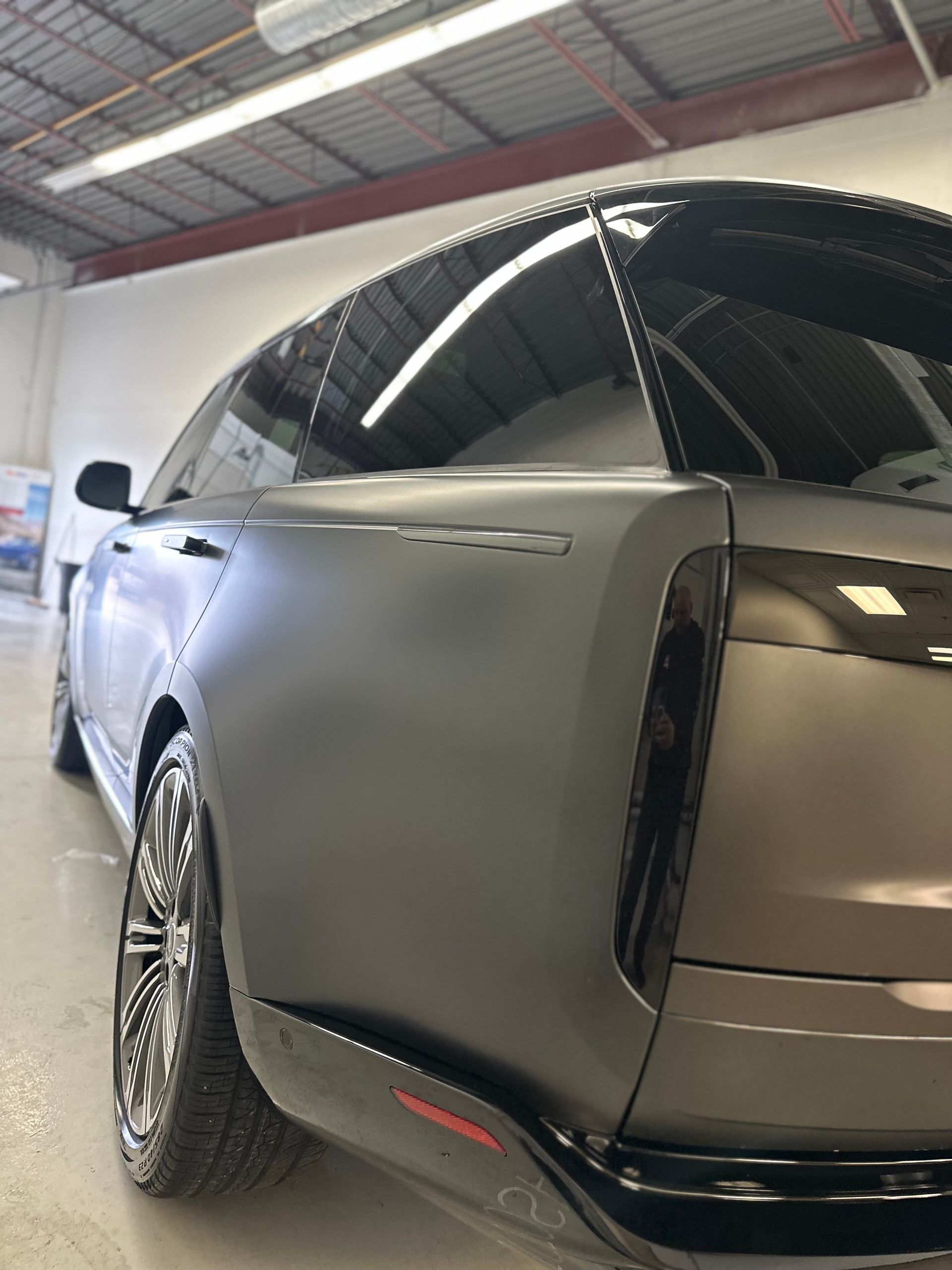 Close-up, low-angle view of the rear quarter of a matte gray SUV in a workshop with black trim and custom wheels.