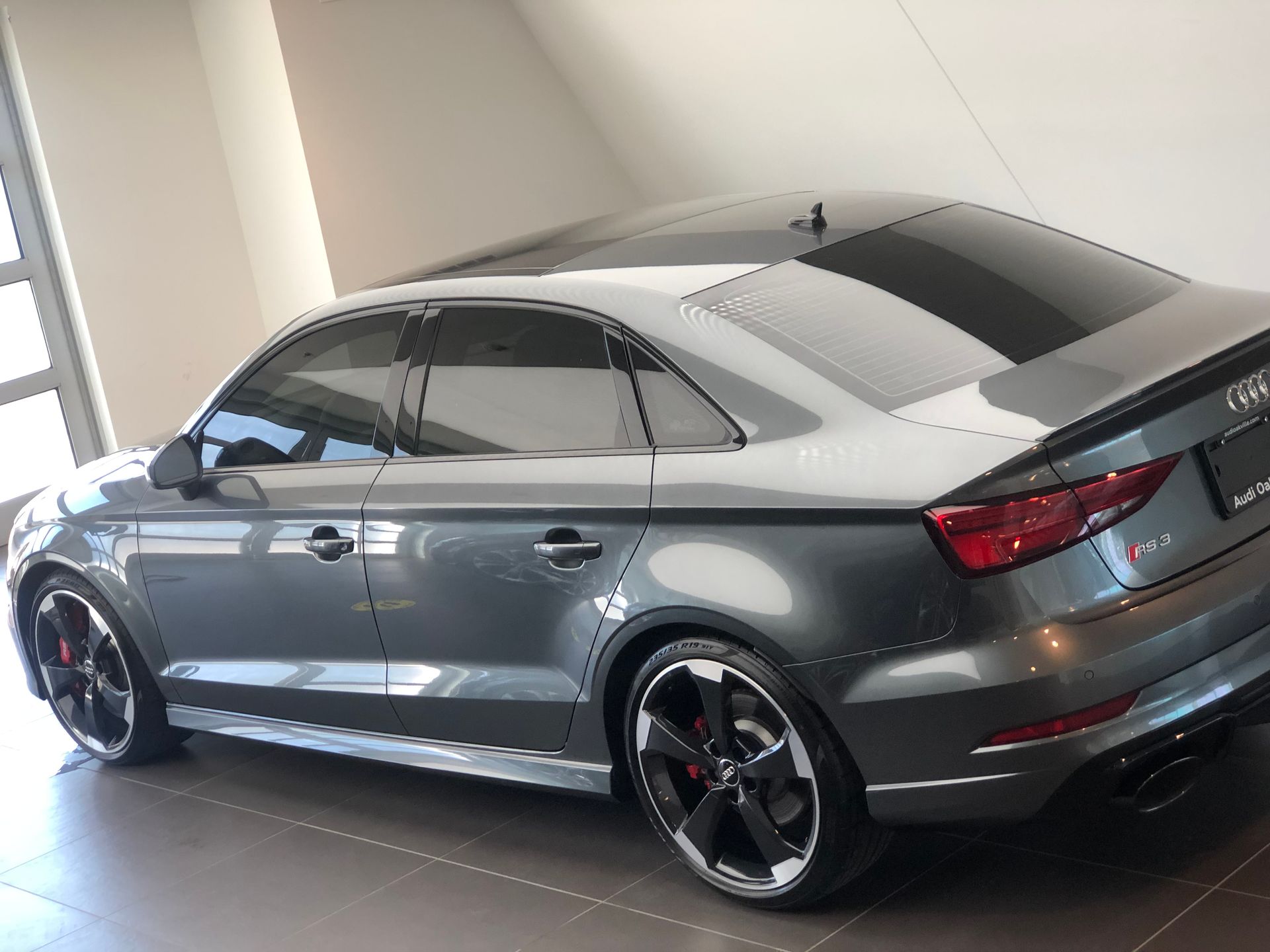 A gray Audi RS 3 sedan parked inside a showroom with tinted windows and black multi-spoke wheels.