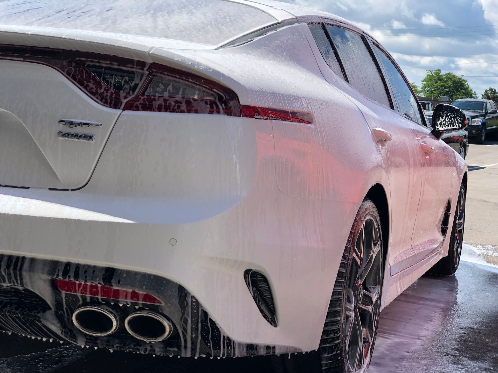 A white Kia Stinger with dual exhaust pipes covered in car wash soap, seen from the rear quarter angle outdoors.