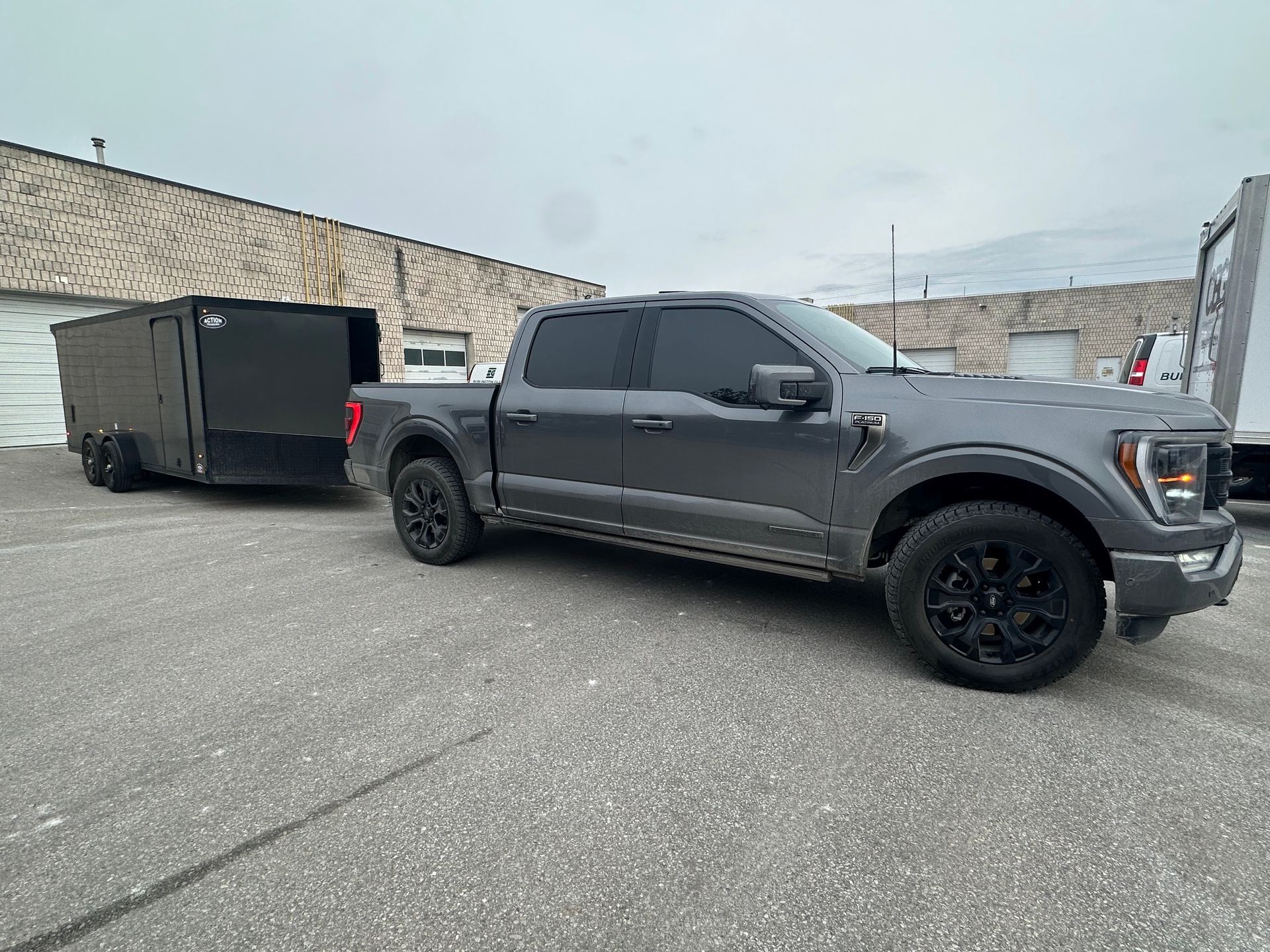 A dark gray Ford F-150 crew cab pickup truck towing a black box trailer parked in a gravel lot against a brick building.