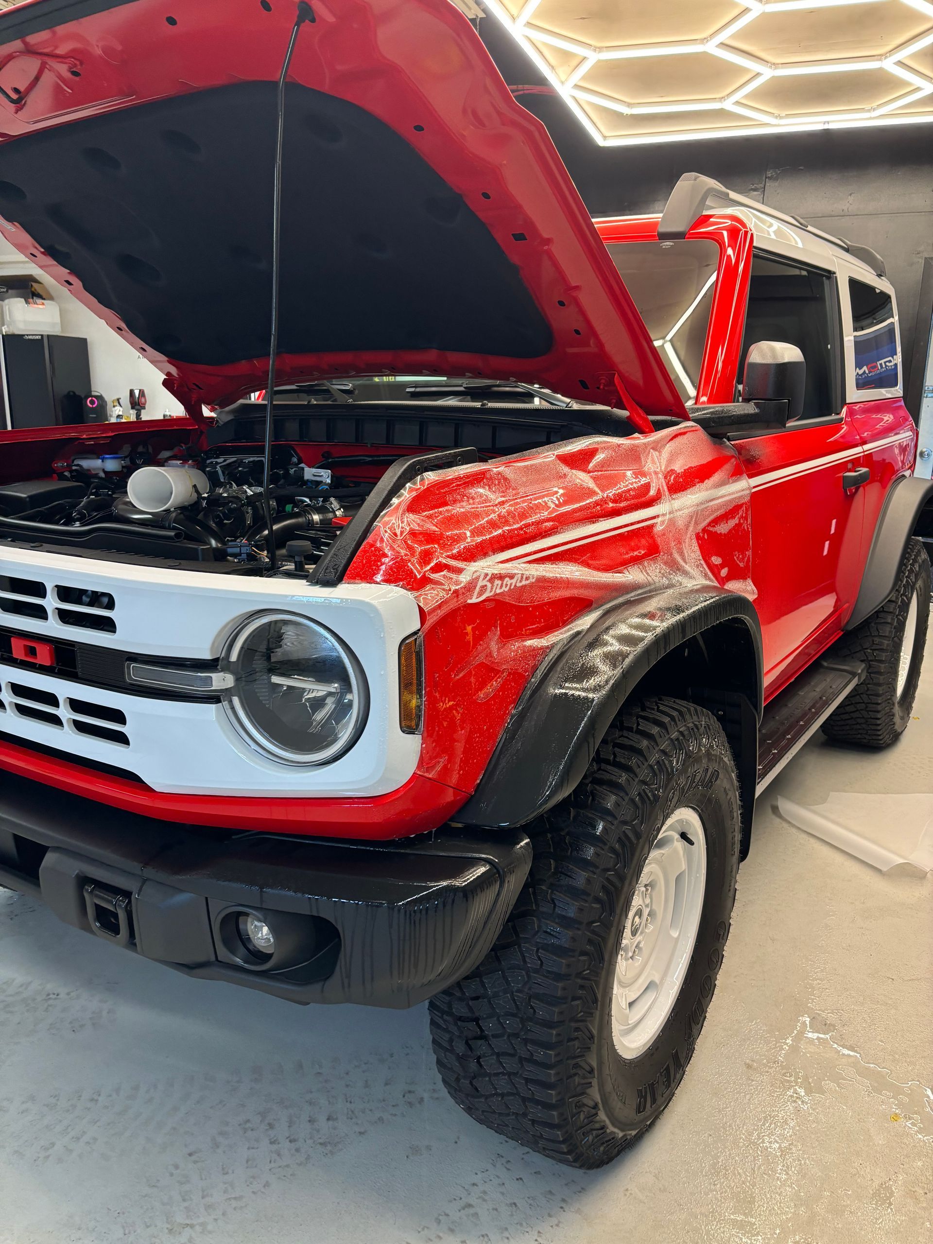 A bright red Ford Bronco with its hood open, parked in a garage with white hex-pattern ceiling lights.