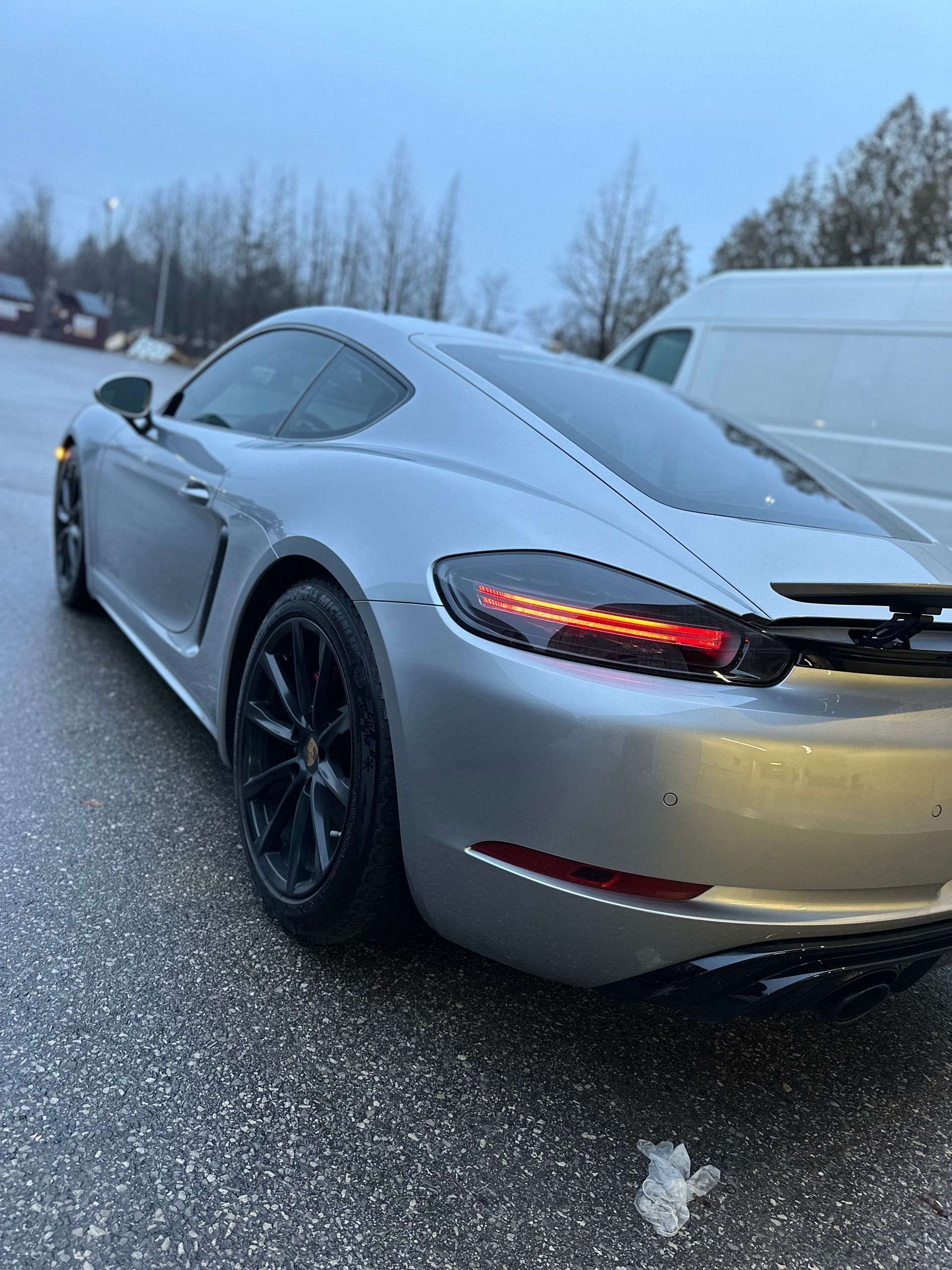 A silver Porsche Cayman sports car parked outdoors on a rainy, overcast day.