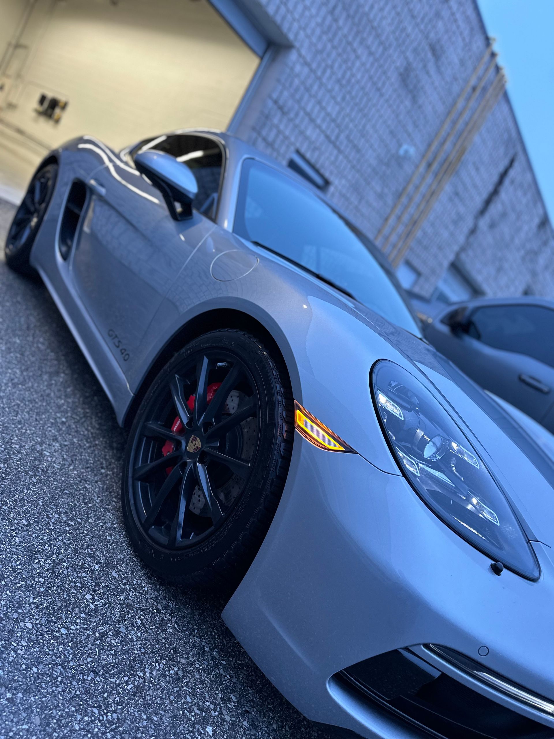 A low-angle, close-up shot of a grey Porsche sports car with black wheels parked outside a warehouse building at dusk.