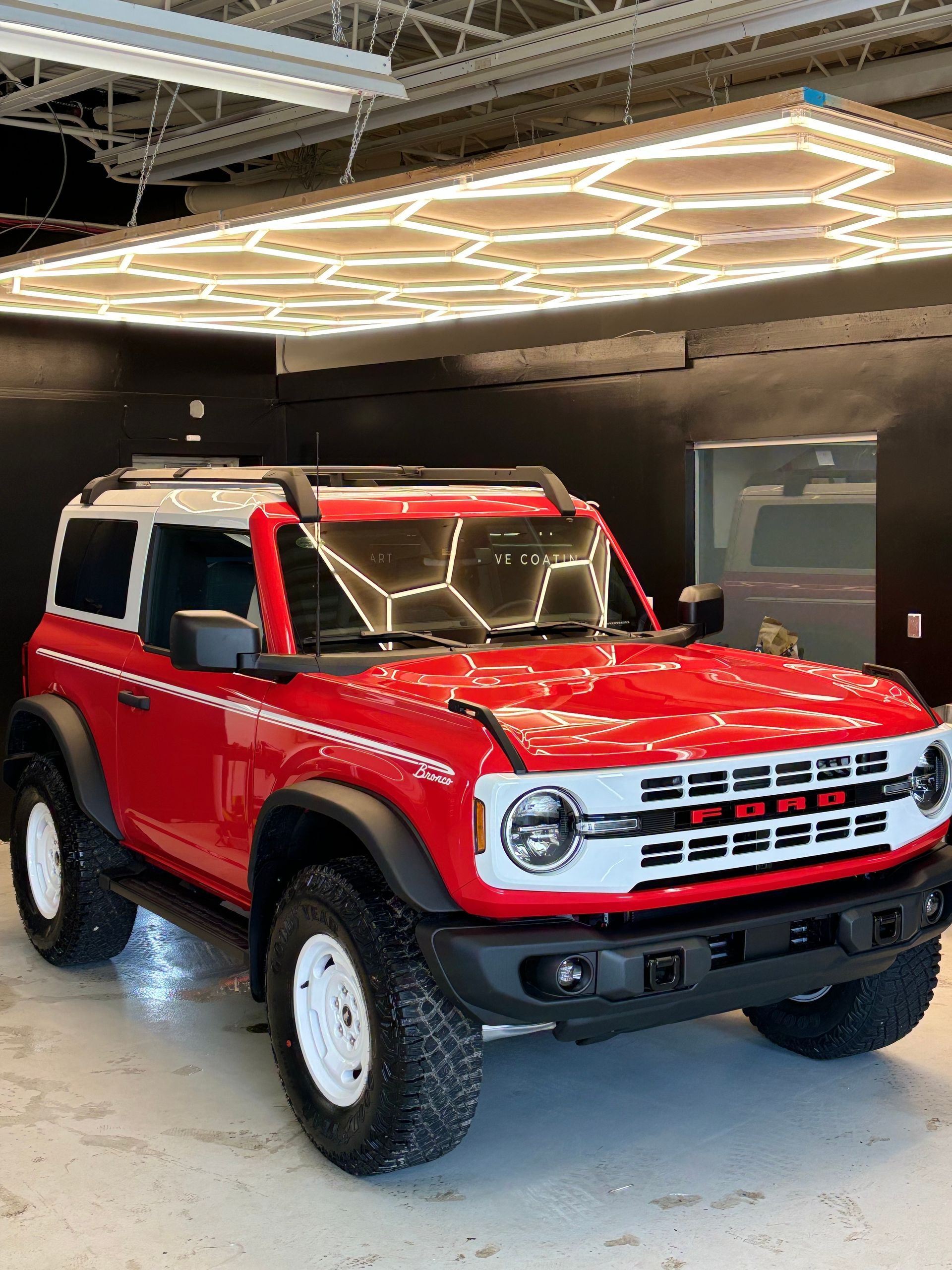 A two-door red and white Ford Bronco parked in a garage with hexagonal overhead lighting.