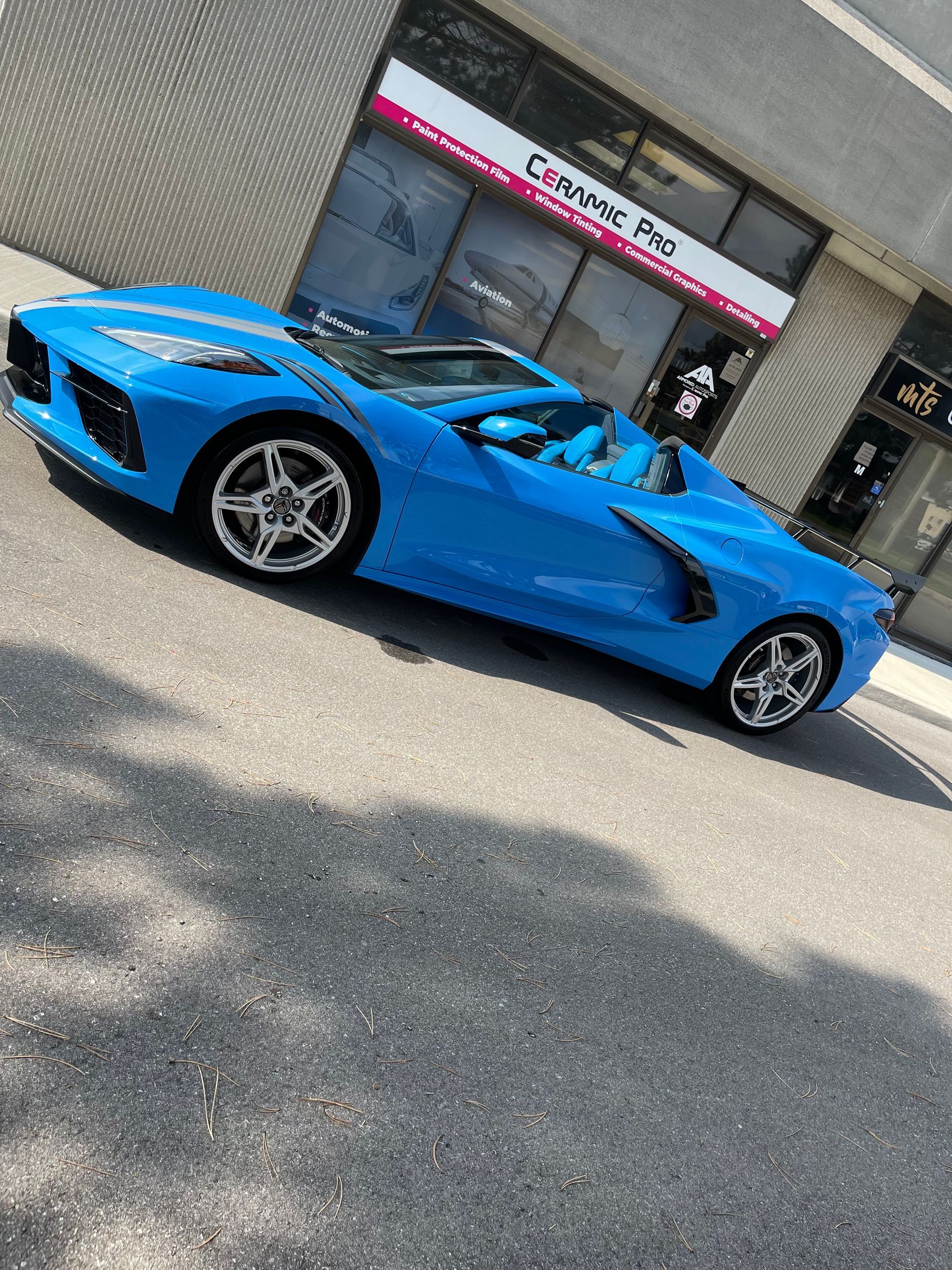 A bright blue Chevrolet Corvette C8 convertible parked on asphalt in front of a Ceramic Pro shop.