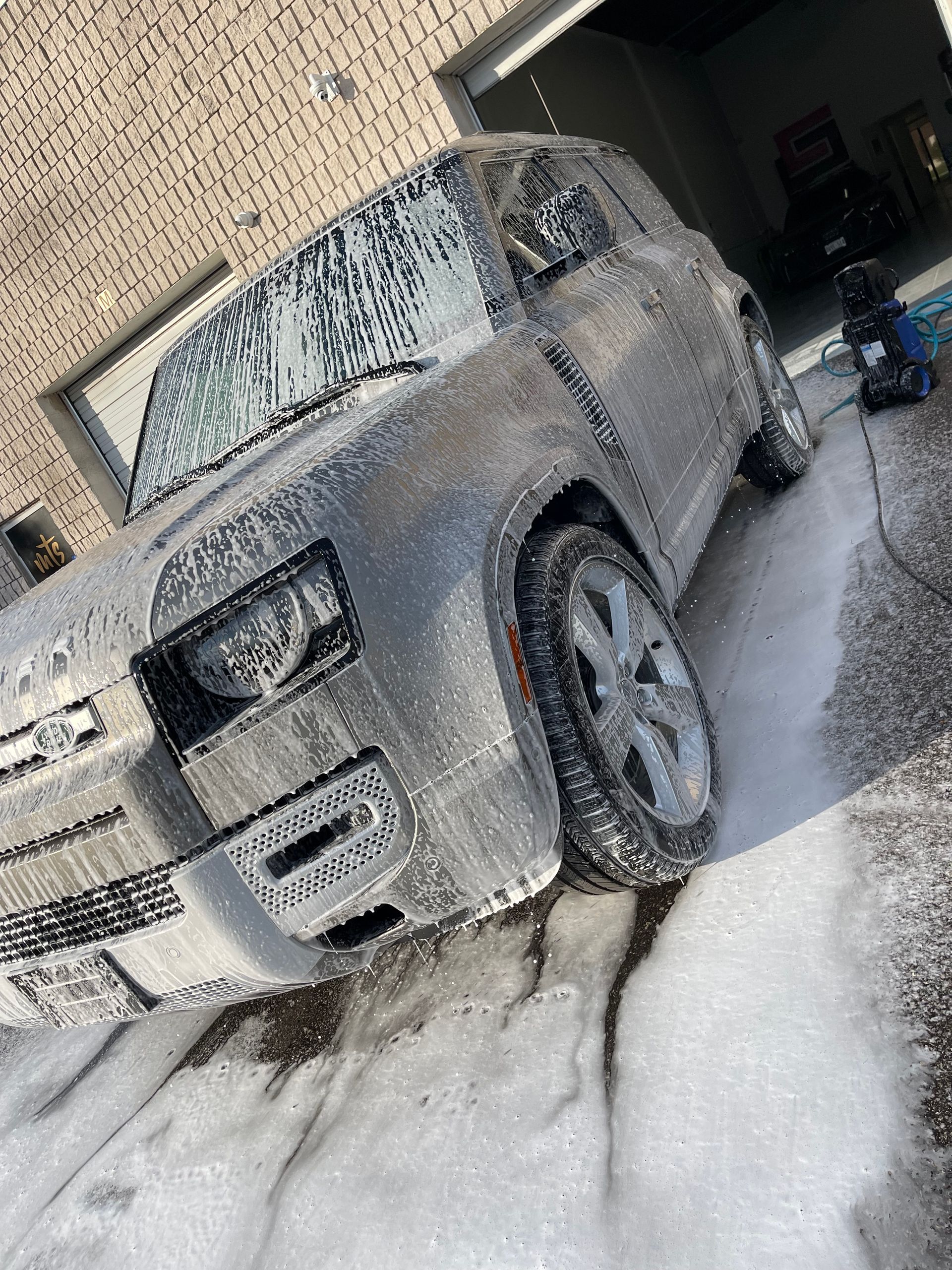 A grey SUV covered in thick white car wash soap, parked outside a garage on a concrete driveway.
