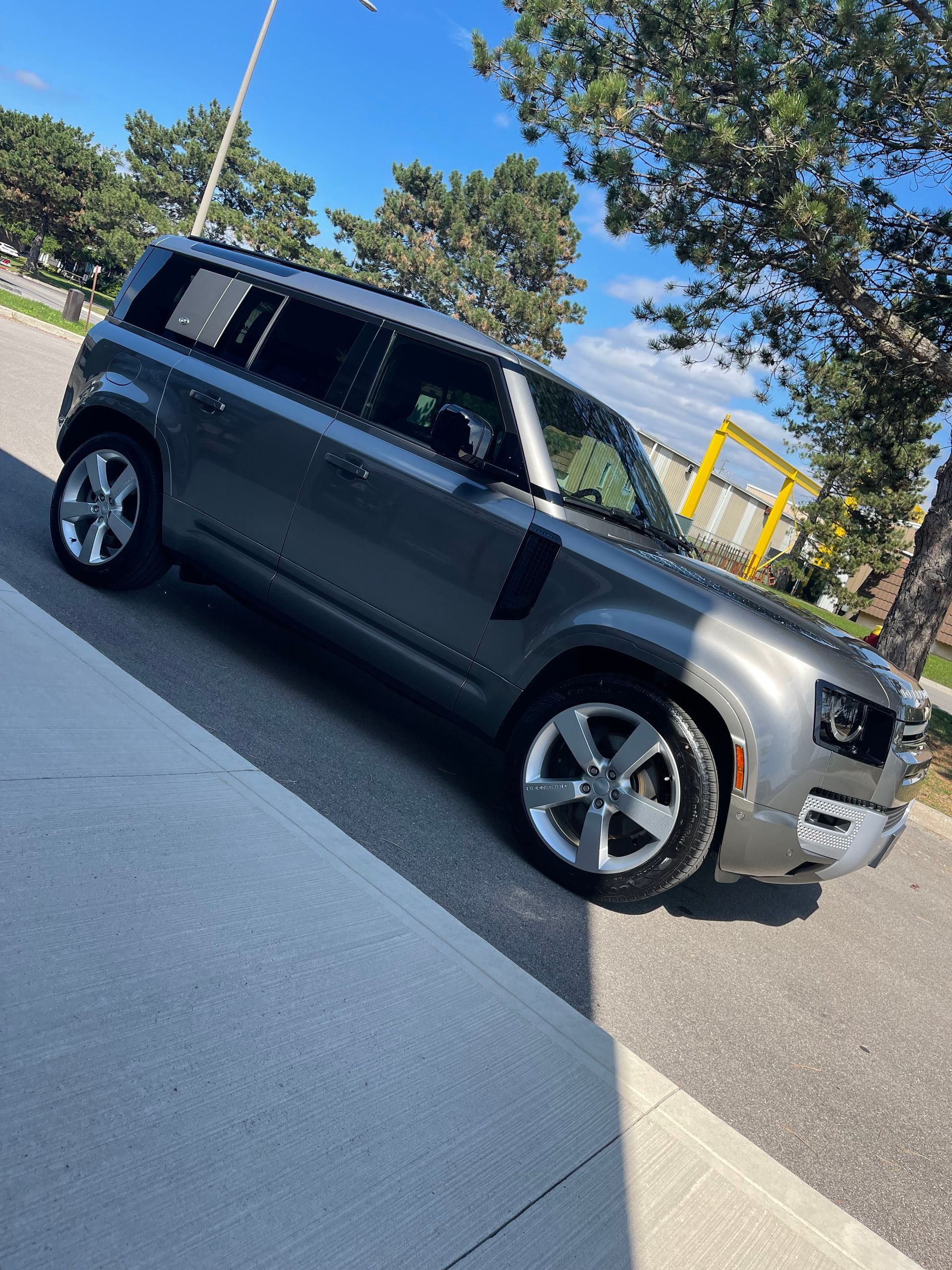 A gray Land Rover Defender parked on a paved lot on a sunny day with trees in the background.