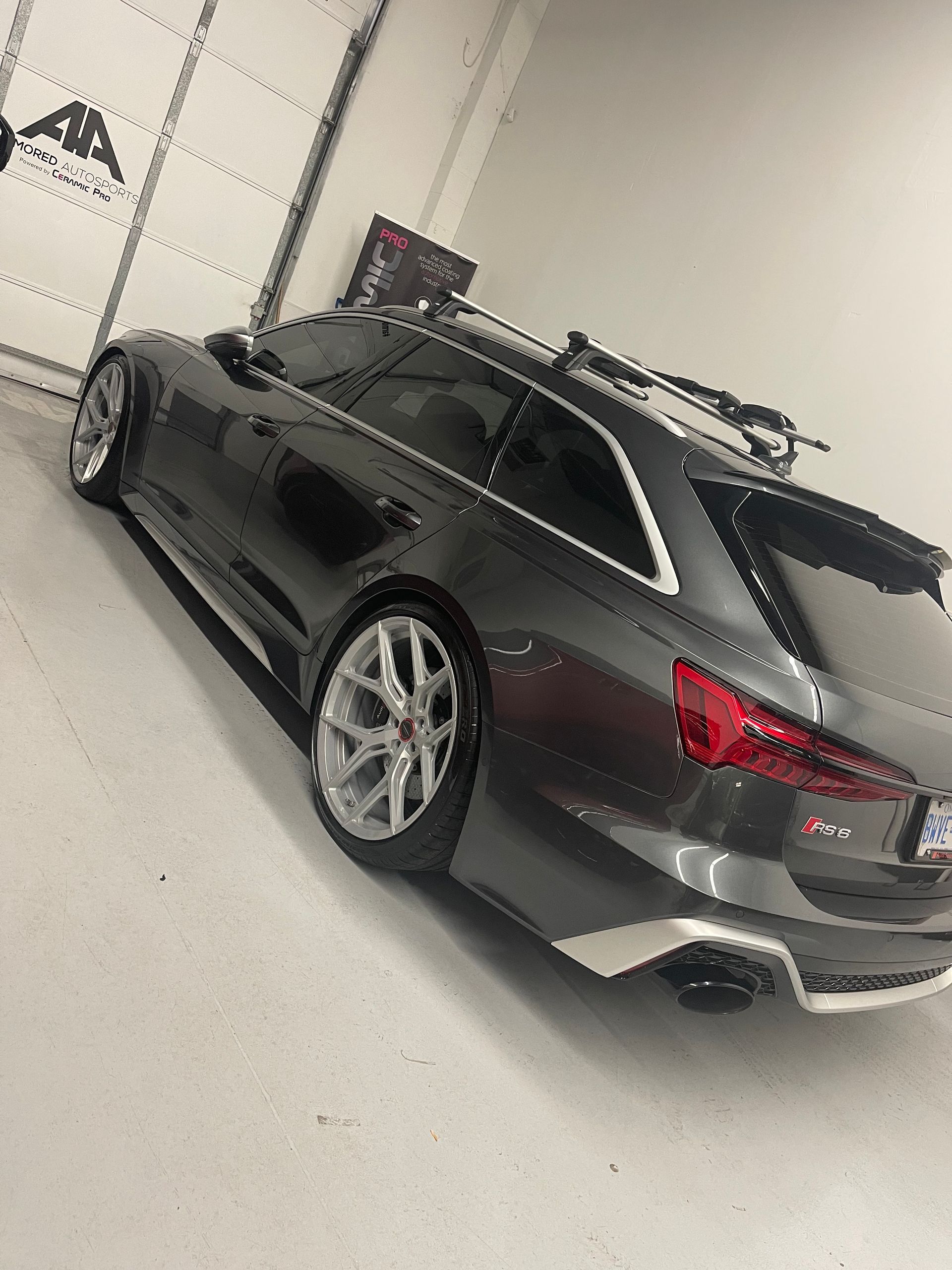 A dark gray Audi RS6 wagon with a roof rack and silver wheels parked inside a white-walled garage.