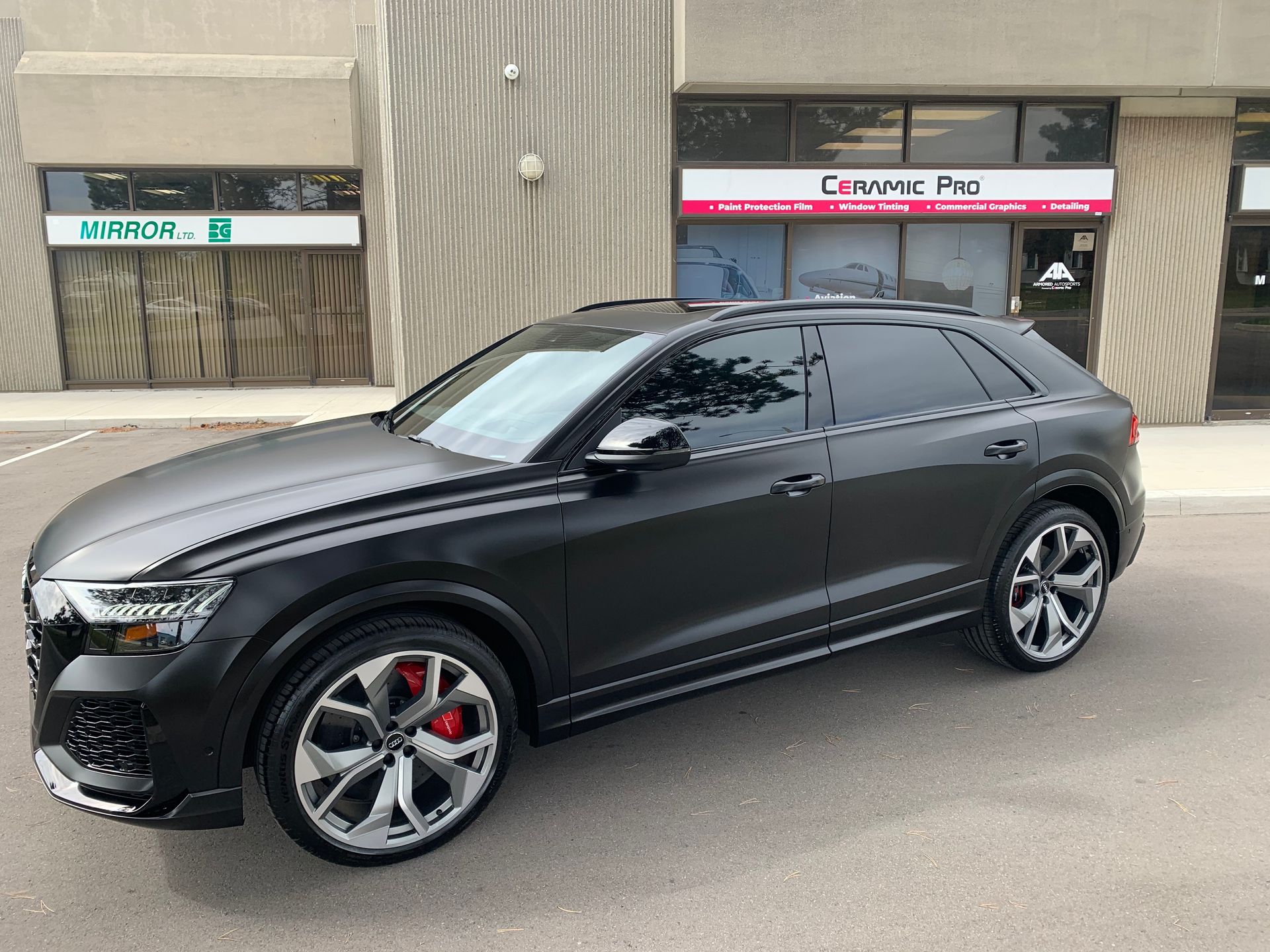 A matte black Audi SUV with large silver wheels and red brake calipers parked in front of a storefront.