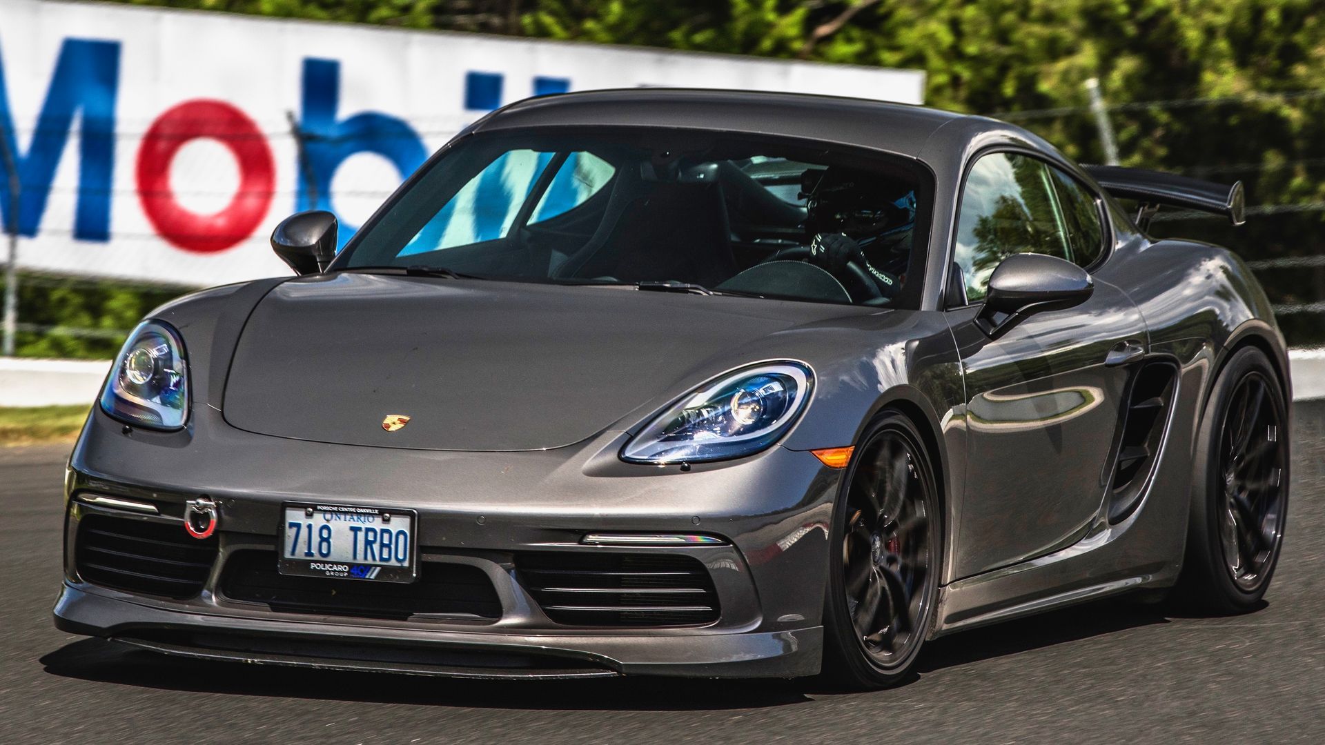 A grey Porsche 718 Cayman GT4 parked on a racetrack in front of a Mobil 1 sign.