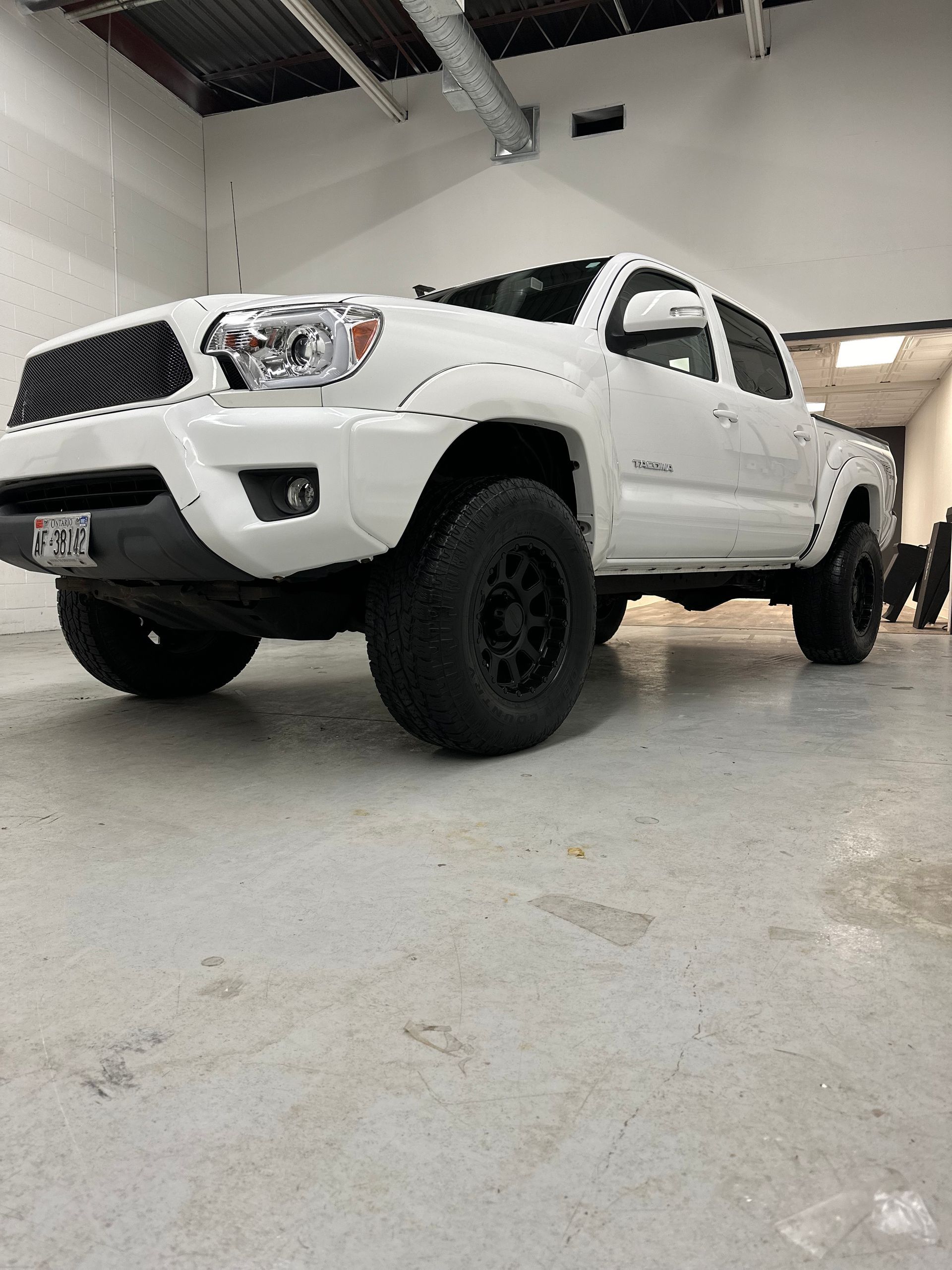 A white Toyota Tacoma pickup truck with black aftermarket wheels and a black grille parked in a warehouse garage.