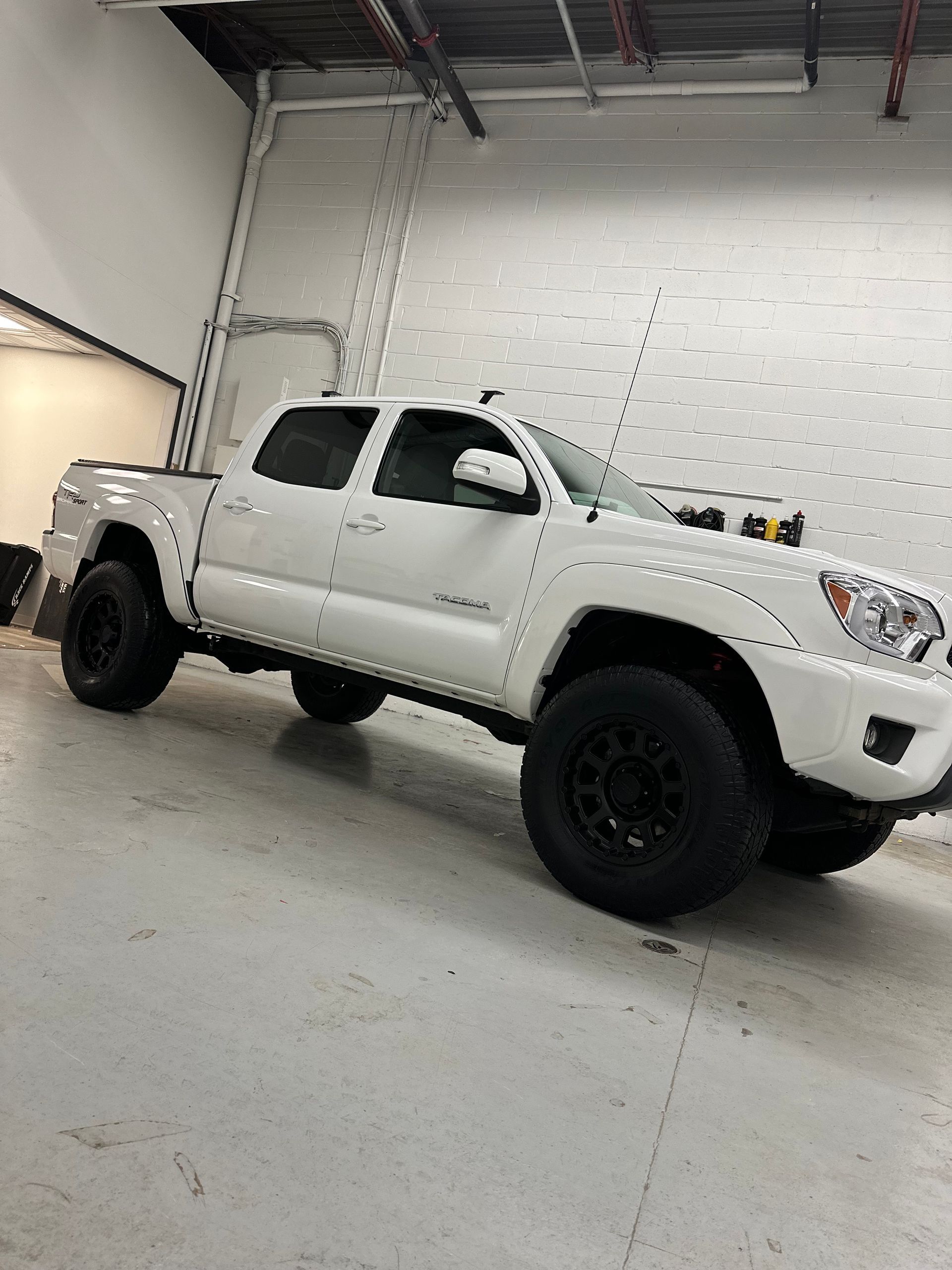 A white Toyota Tacoma pickup truck with black aftermarket wheels and tires parked in a brightly lit indoor warehouse.