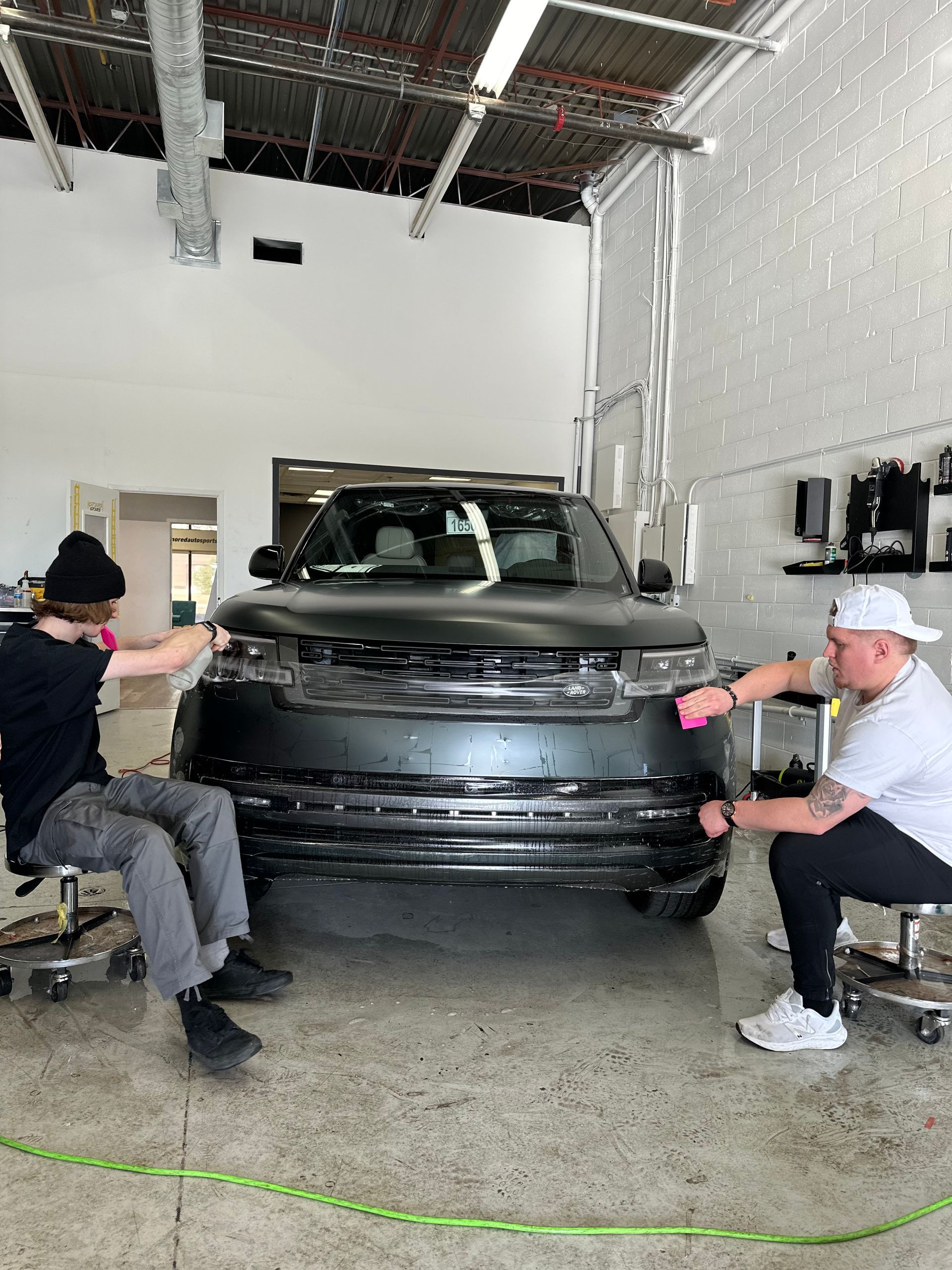 Two technicians in a garage work on the front bumper of a black SUV.
