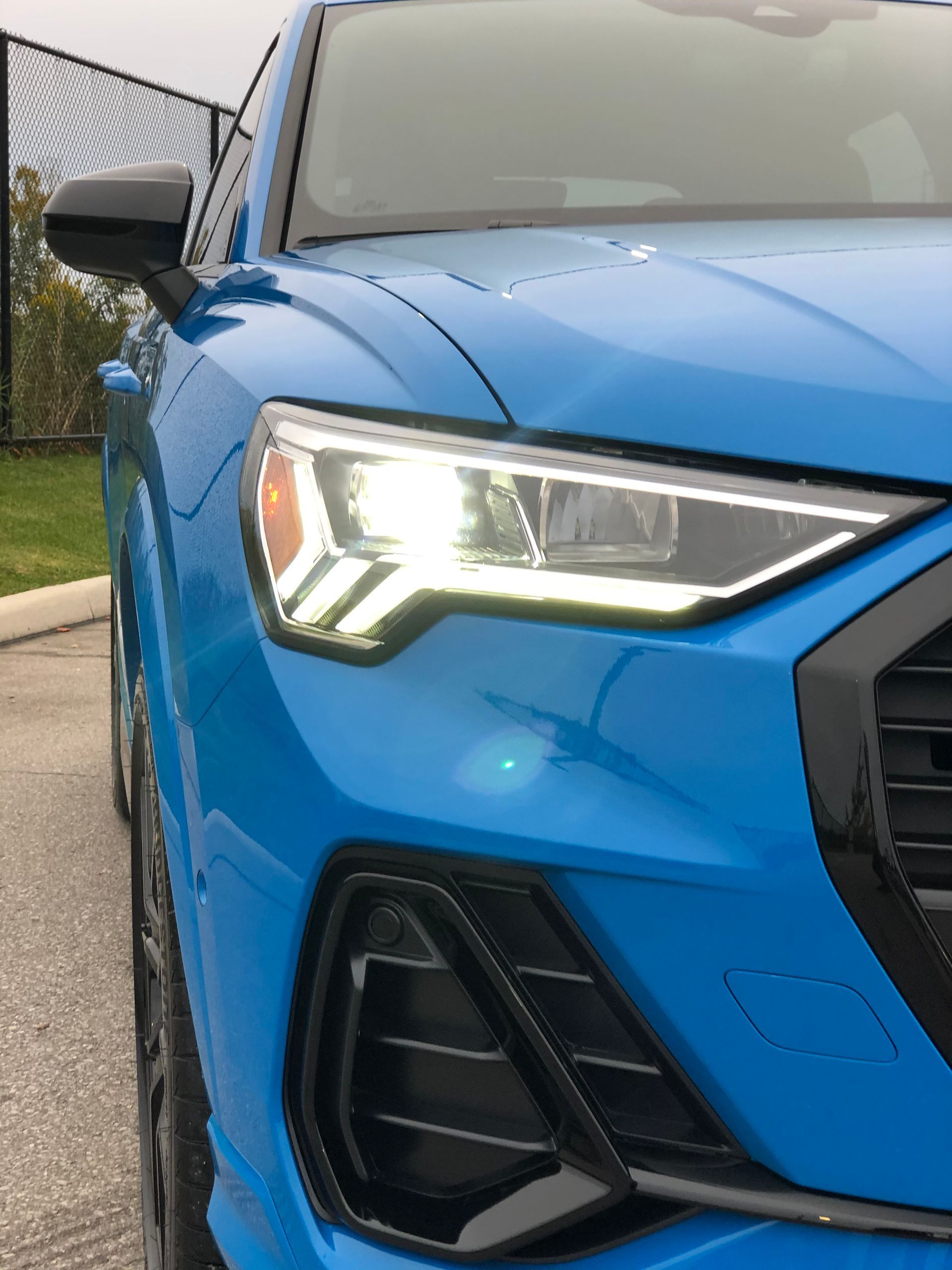Close-up of a bright blue Audi SUV's front headlight, grill, and air intake on a paved surface.