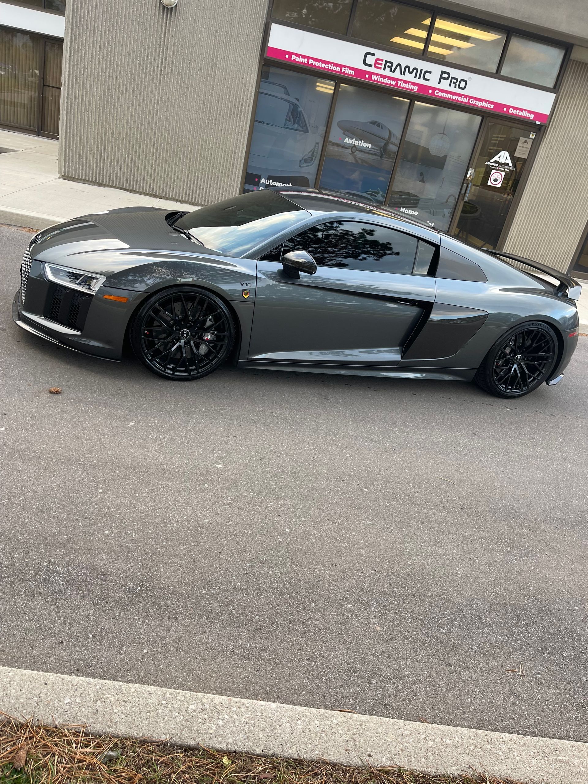 A dark gray Audi R8 parked on asphalt in front of a storefront.