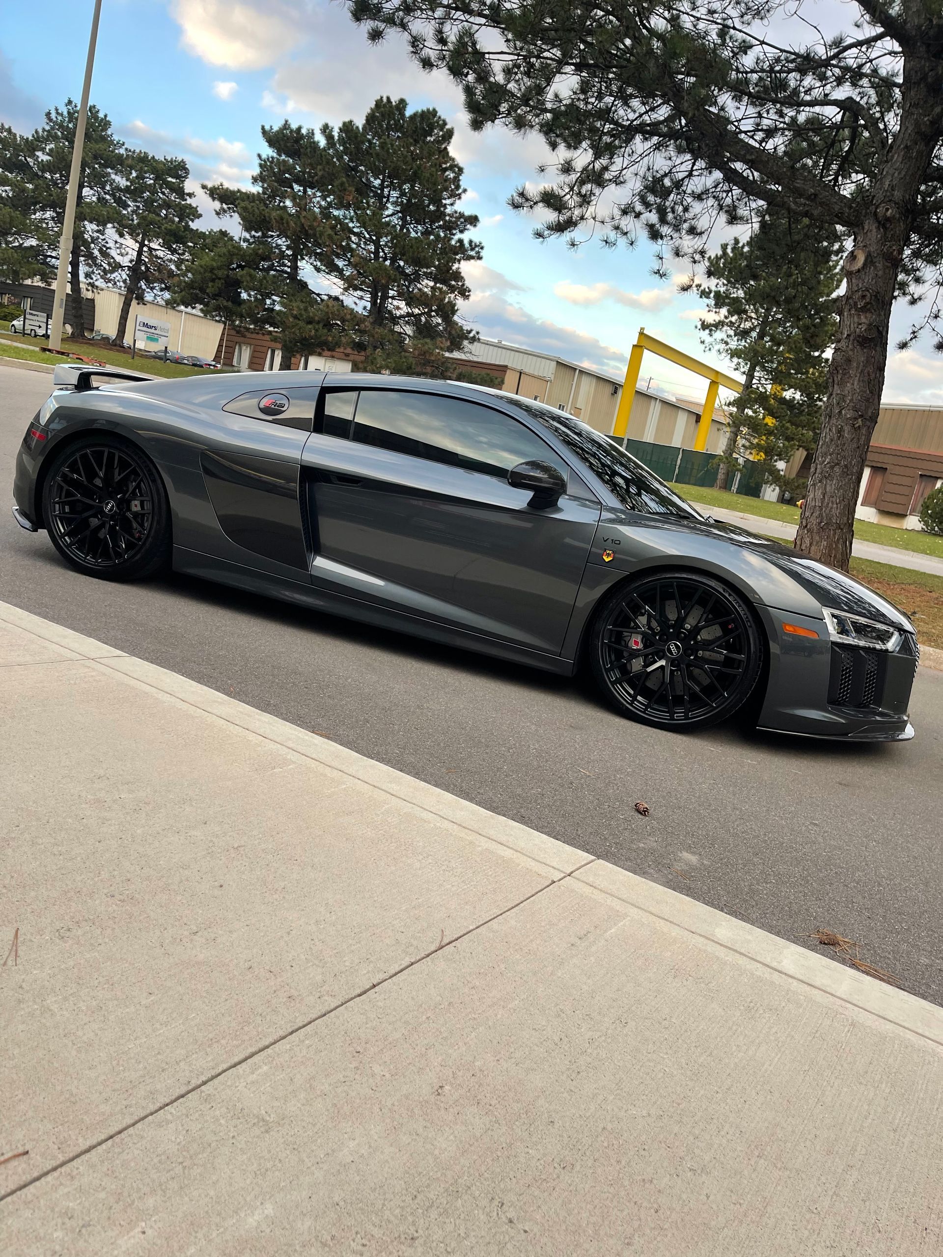A sleek, dark gray Audi R8 sports car parked on the edge of a paved road with trees in the background.