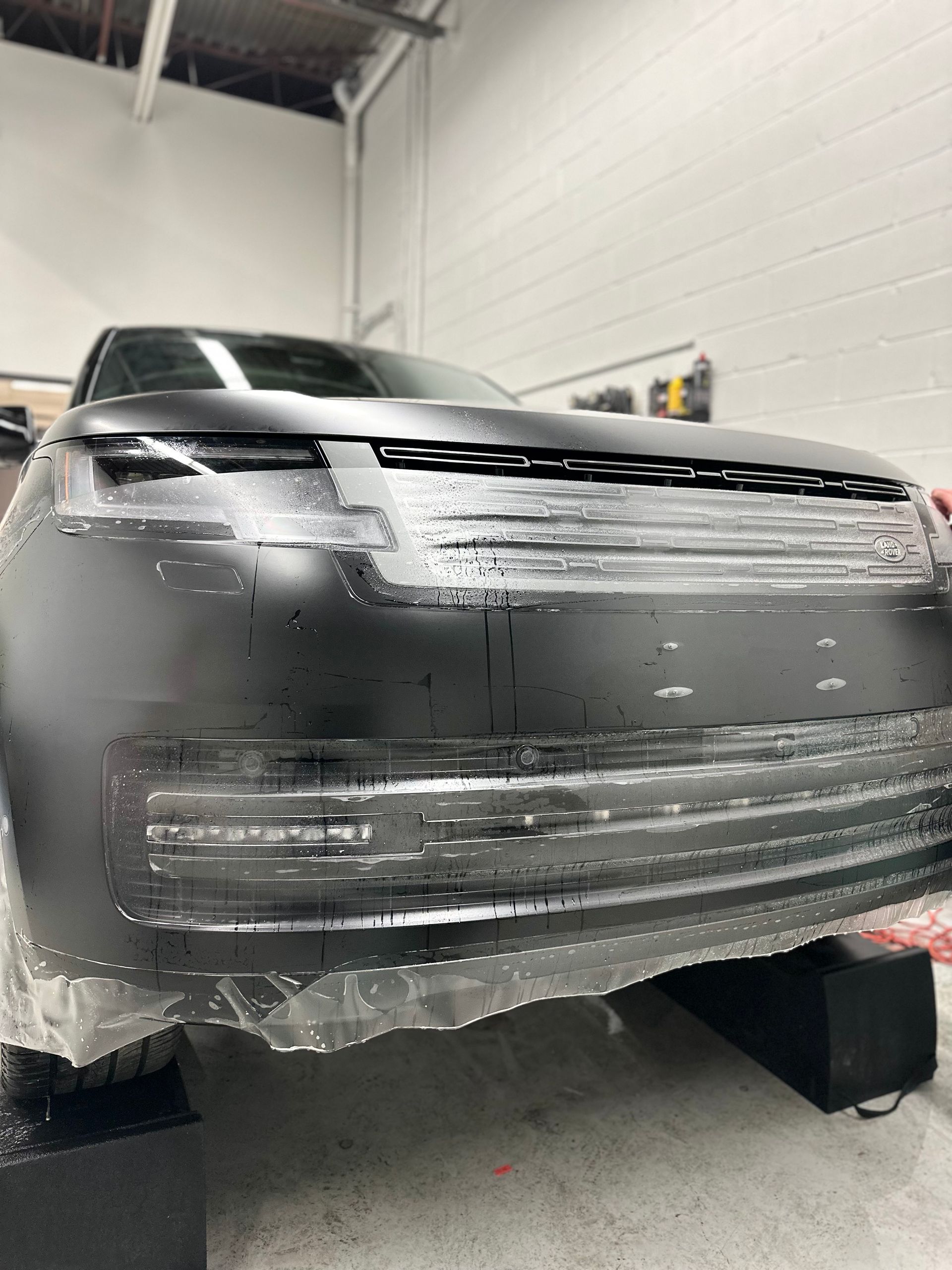 A matte black Range Rover being fitted with protective film in a garage, with clear film strips visible over the grille.
