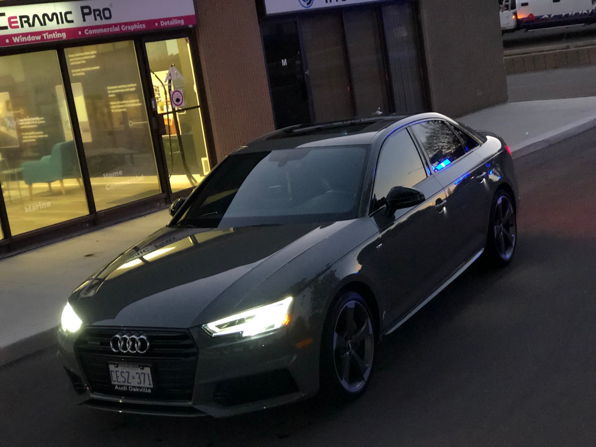 A gray Audi sedan parked in front of a Ceramic Pro shop, illuminated by its bright headlights at dusk.