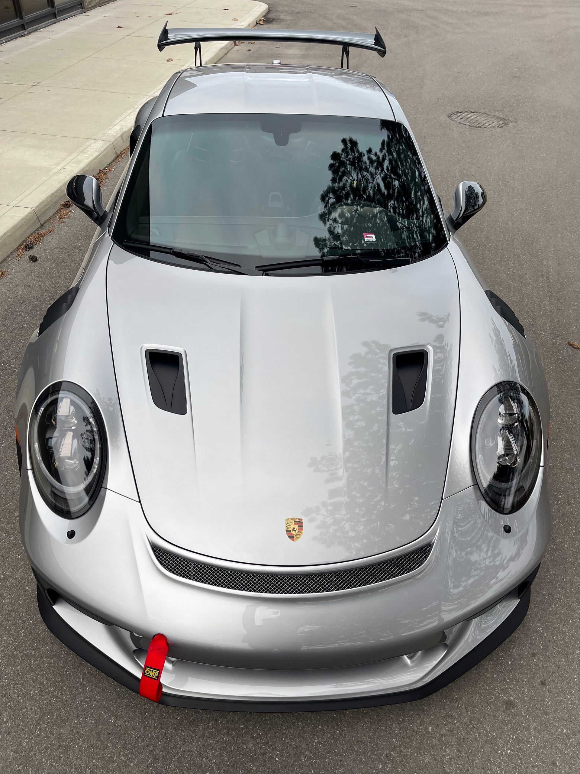High-angle front view of a silver Porsche sports car with a rear wing and red tow strap, parked on pavement.